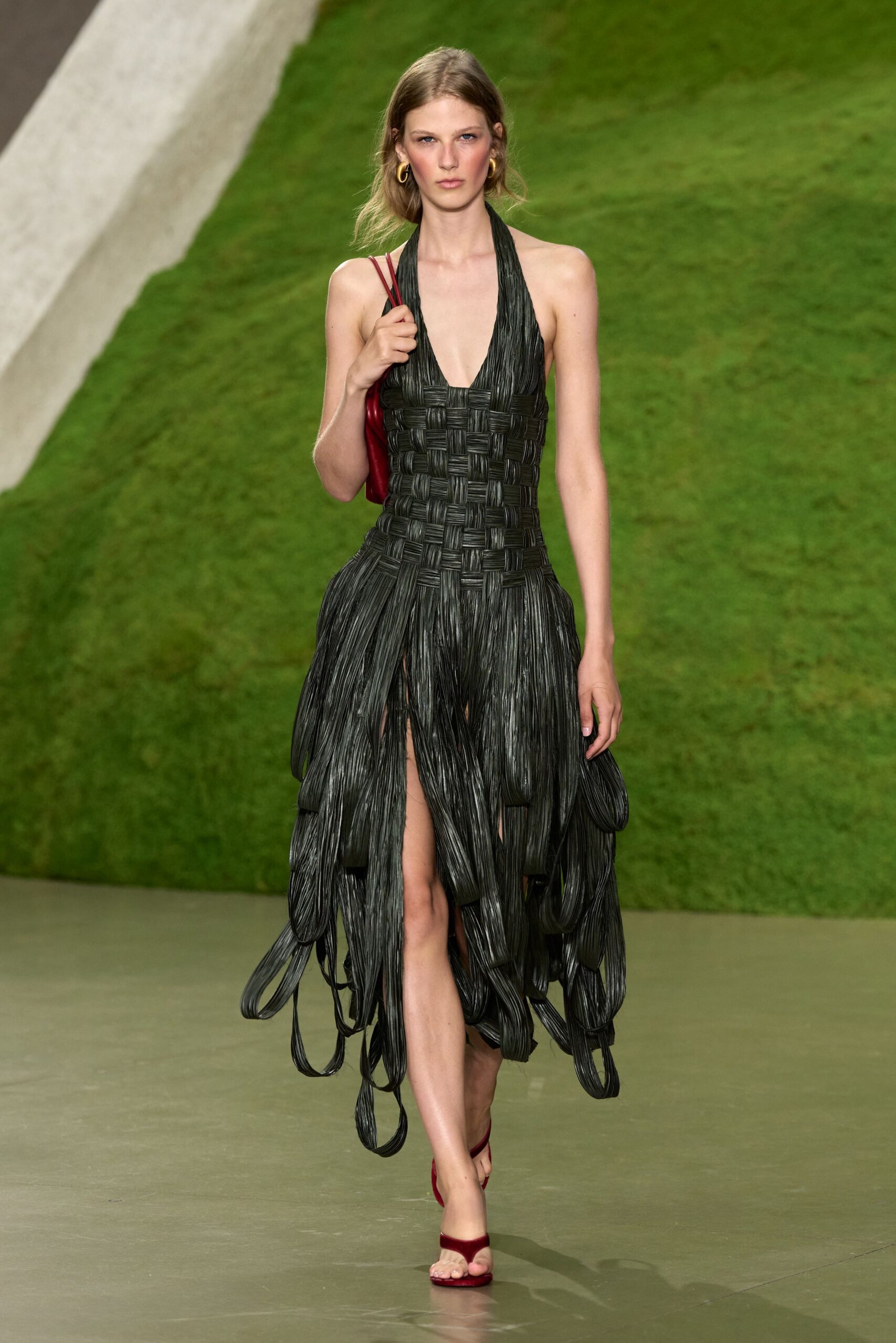 Chunky gold hoops. Black halter dress woven in broad glossy bands, looped lengths swinging below the knee. Small oxblood shoulder bag tucked high under the arm. Red velvet thong sandals, barely there at the toe. Crossing a sculpture garden after rain, invitation folded in the bag.