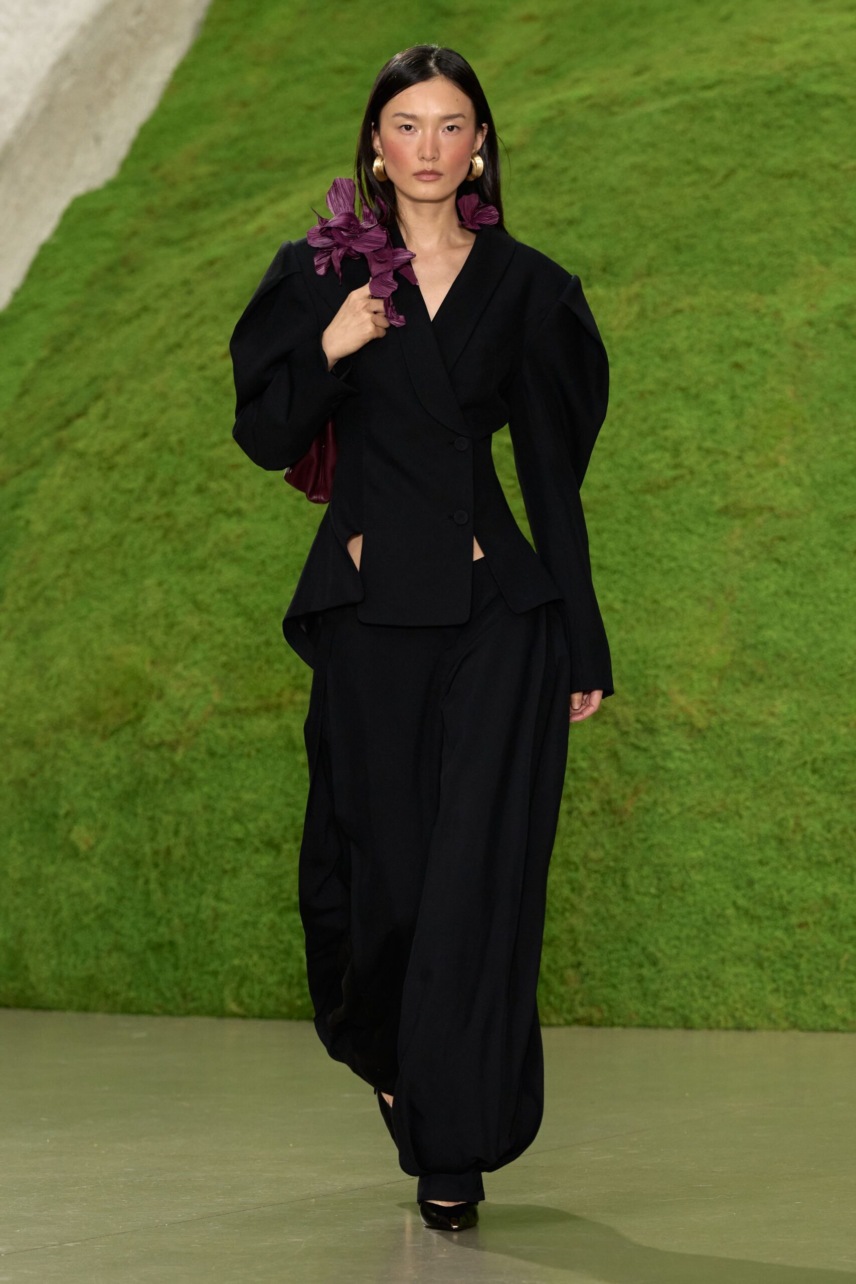 Large gold hoops, thick as door knockers. Black tailored blazer with puffed sleeves and waist cutouts. Burgundy shoulder bag wrapped in pleated orchid sprigs. Black trousers falling loose into a soft pooled hem. Pointed black shoes with a glinting metal toe. Crossing a sculpture garden after rain, invitation folded in your fist.