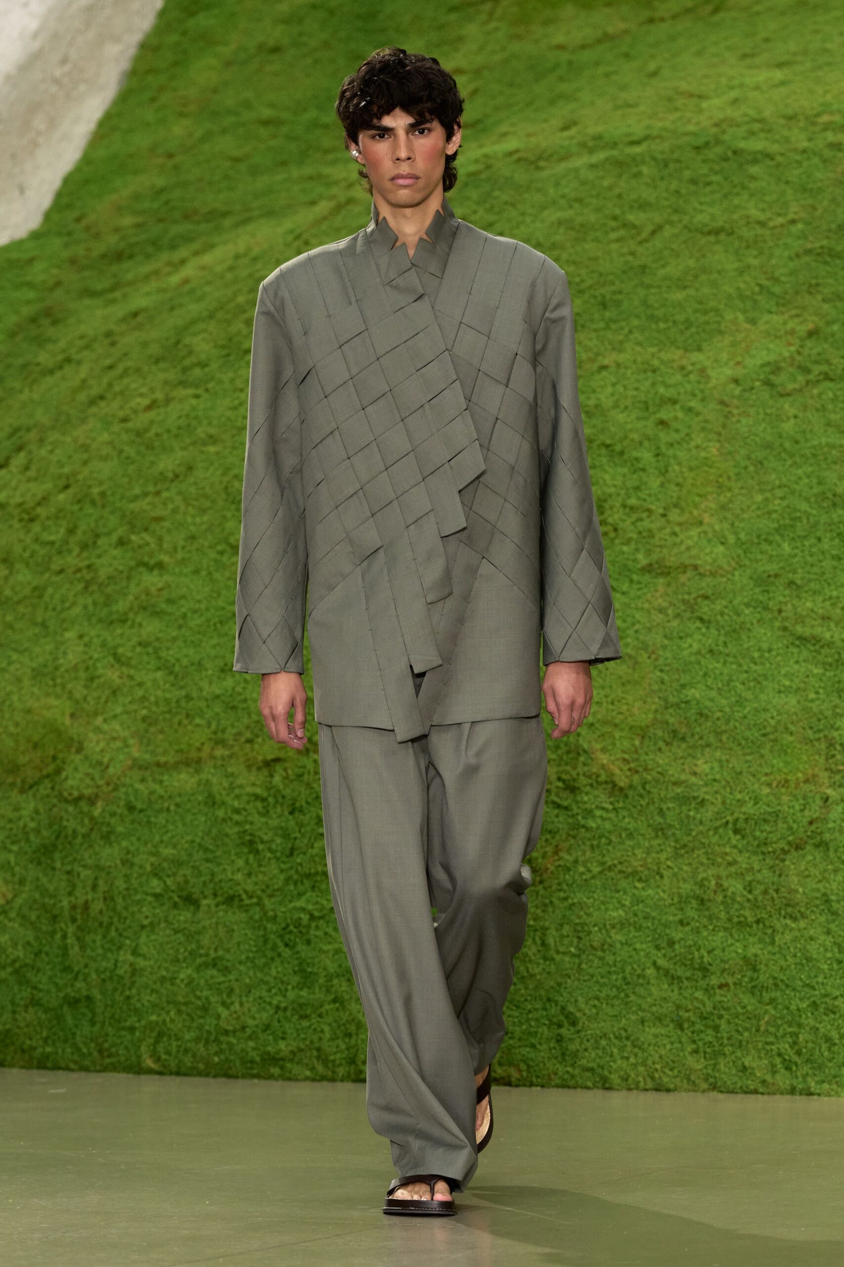 Sage tailoring woven into broad lattice bands, one strip hanging longest. Loose straight trousers in the same fine suiting cloth. Flat black thong sandals. Crossing a lawn after the vows, jacket still half unspooling.