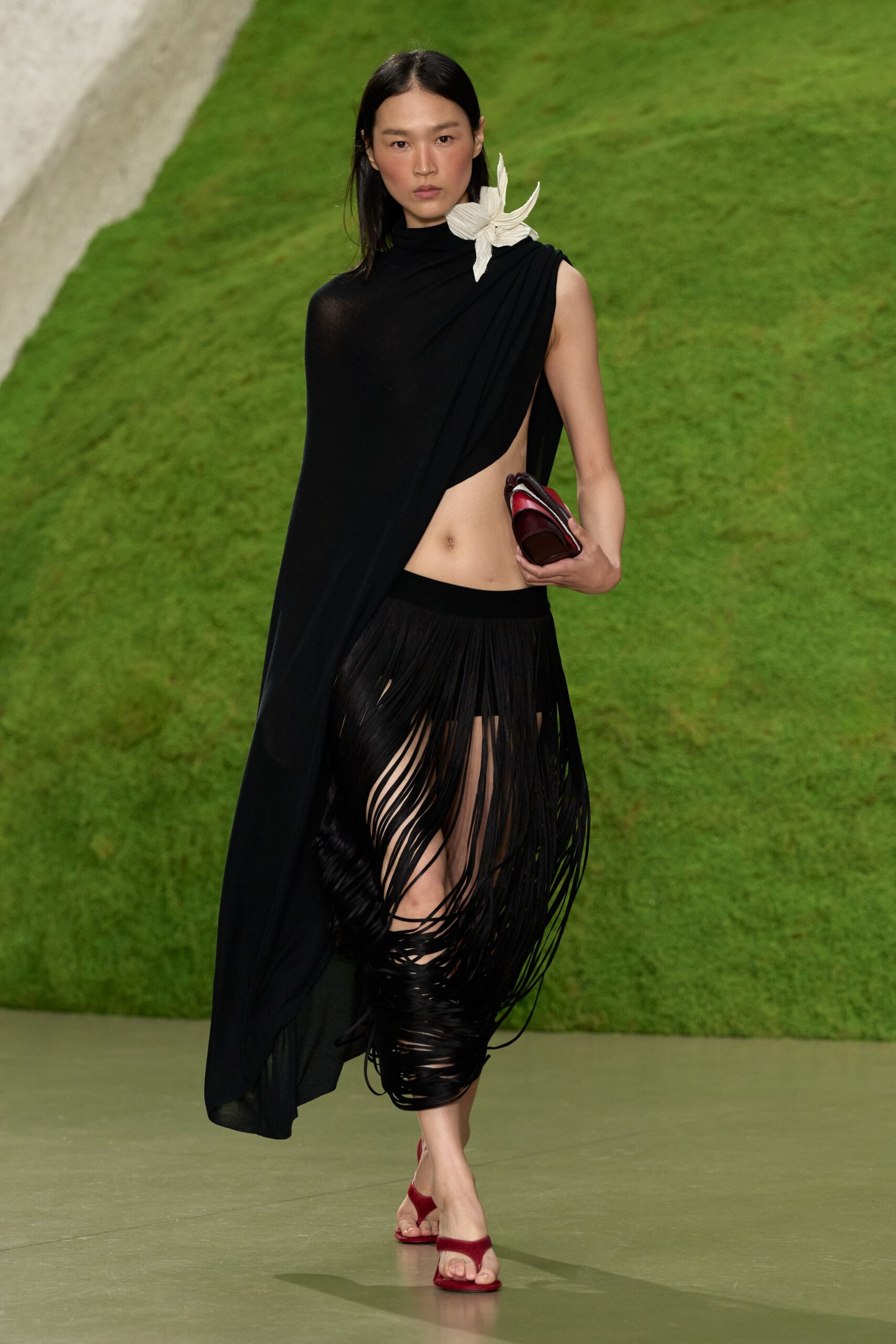 Black jersey column draped one-shoulder, one side cut to the hip. Pleated ivory petals pinned high at the collarbone. Dense black strands swish from a narrow waistband to mid-calf. Burgundy folded clutch tucked under the arm. Wine-red velvet thong sandals. Crossing a lawn after dark with your shoes in danger from the dew.