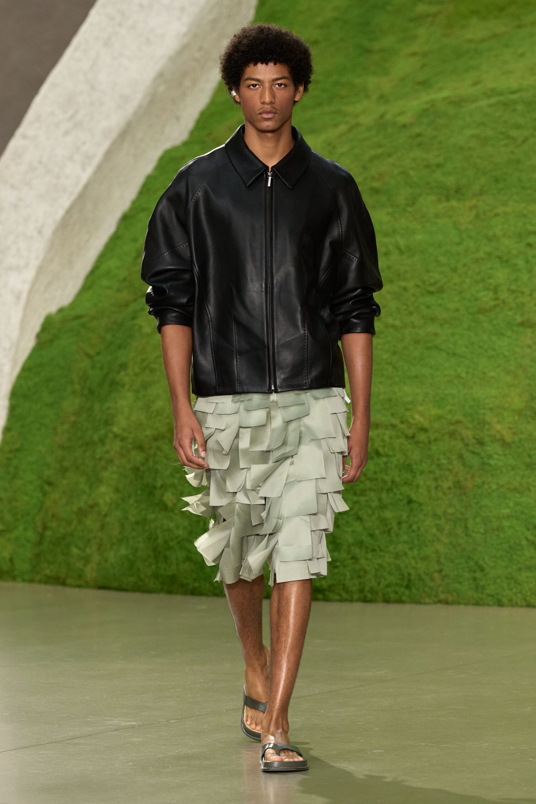 Black leather zip jacket with ballooned sleeves and curved paneled seams. Pale sage shorts buried under overlapping organza tabs like clipped leaves. Black leather thong slides with a squared foam sole. Walking from a greenhouse dinner onto wet paving after the sprinklers ran.