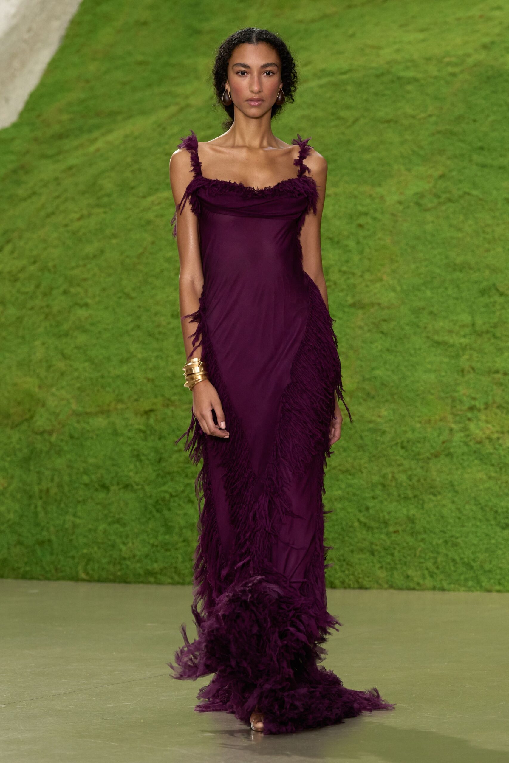 Large bronze hoop earrings. Plum column gown in clingy jersey, feathered at straps, hips and sweeping hem. Stacked gold cuffs with jagged ridges. Barely there metallic sandals. Crossing a lawn after midnight with damp grass catching the train.