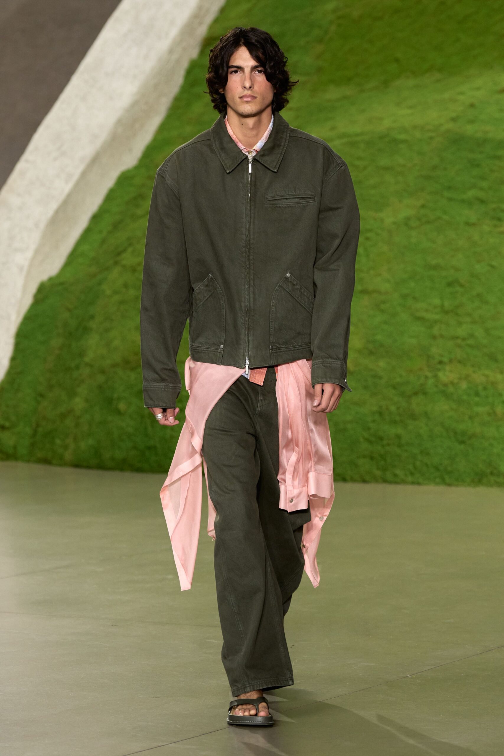 Washed olive zip jacket with curved welt pockets and a bright metal pull. Pale pink shirt hanging untucked below the hem, tails flapping open. Matching olive denim trousers cut loose through the leg. Black leather thong sandals with a chunky sole. Walking back from the beach road dinner before the air turns cold.