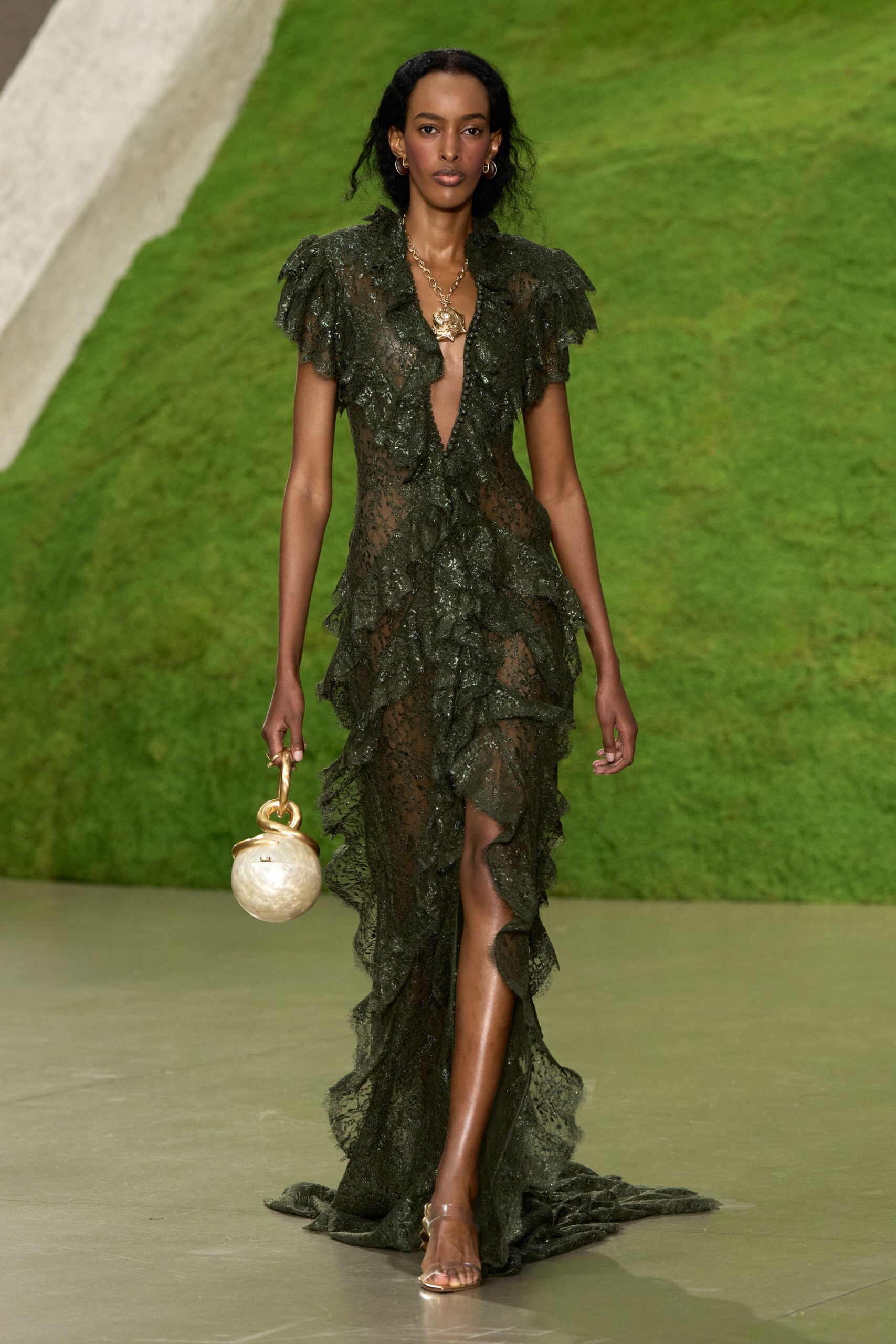 Small gold hoops hugging the lobe. Heavy gold chain with a shell-like pendant at the plunge. Sheer swamp-green lace gown with ragged ruffles and a thigh-high slit. Round mother-of-pearl sphere bag with a thick gilt loop handle. Barely there metallic sandals. Walking back from a beach wedding after the sky turns storm-green.