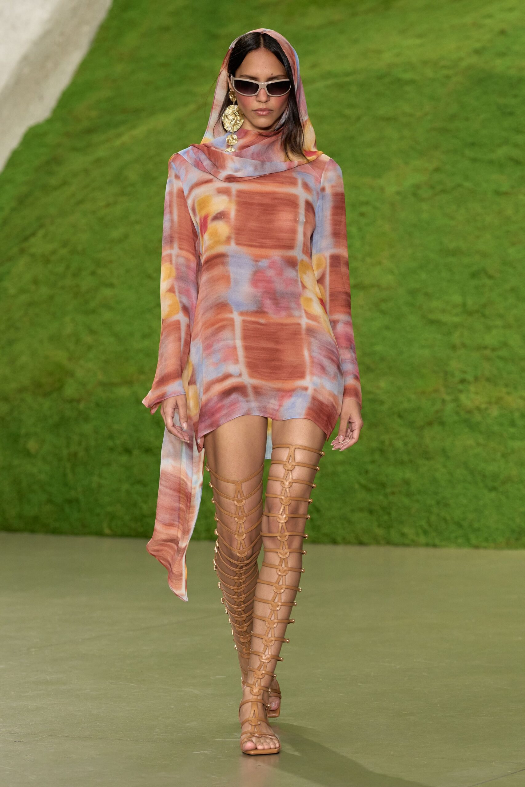 Narrow silver-rim sunglasses with smoked lenses. Hammered gold disc earring with smaller medallions swinging below. Sheer mesh mini in sun-faded rust checks, scarf hood draped long at one side. Tan leather cage sandals climbing the thigh with ring-linked straps. Walking back from a beach club after the sun has gone soft.