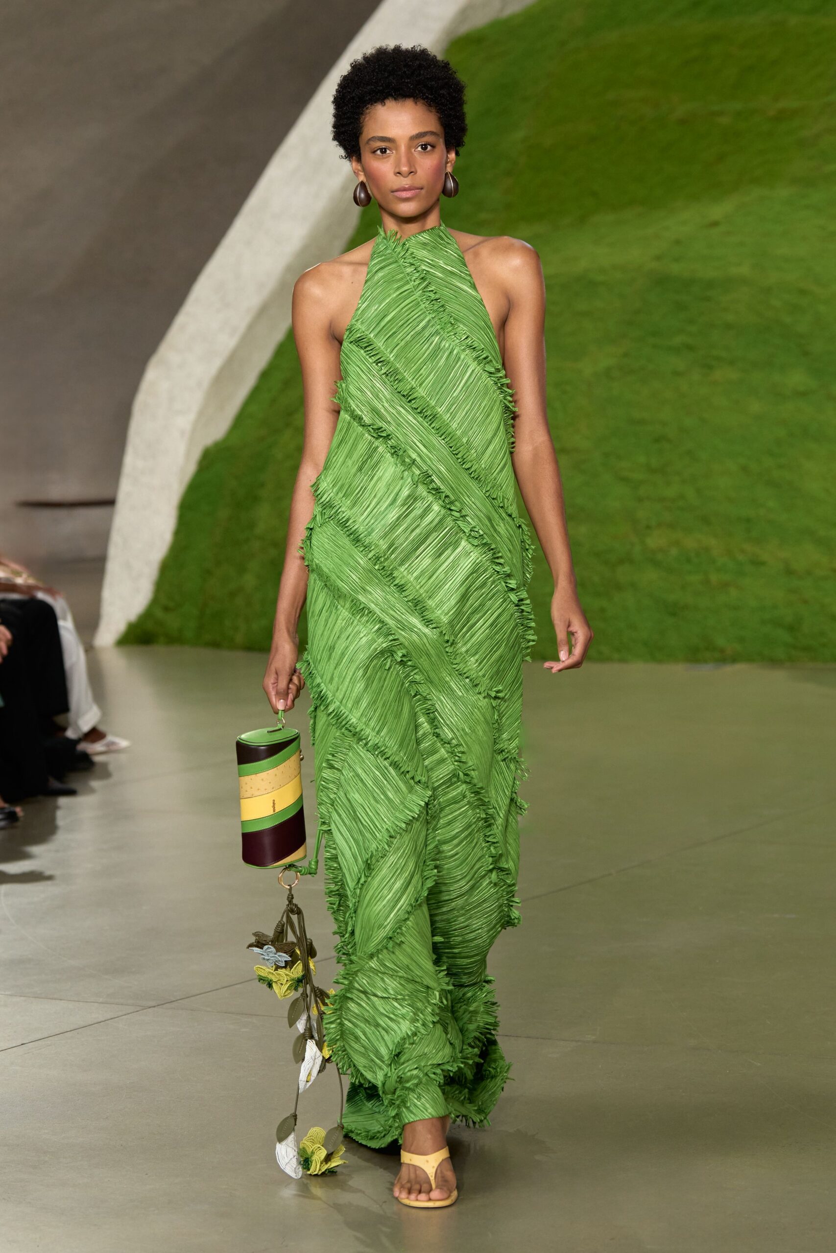 Dark wood drop earrings, smooth and egg-shaped. Grass-green halter column gown built from diagonally pleated panels with fringed joins. Cylindrical colour-block bag with a vine of crocheted leaves and flowers. Butter-yellow thong sandals. Walking from a conservatory lunch into heat that hits like wet glass.