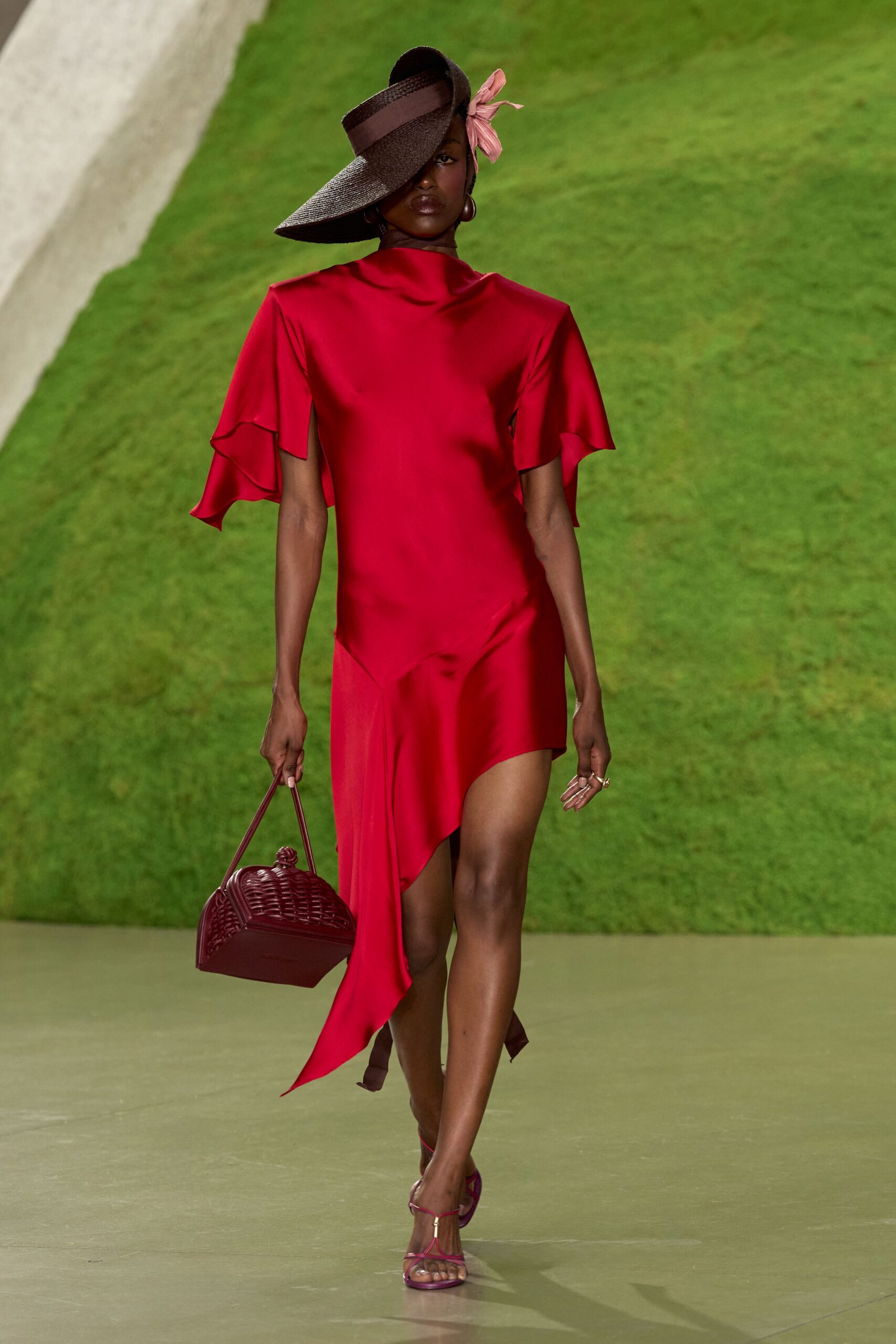 Wide chocolate straw visor, tilted low with a dusty-rose bow. Crimson satin dress with split flutter sleeves and a handkerchief drop hem. Large oxblood drop earrings. Burgundy wicker top-handle bag with a knotted lid. Berry T-strap sandals, slim metal bar at the toe. Garden wedding heat, grass still damp where the chairs were.