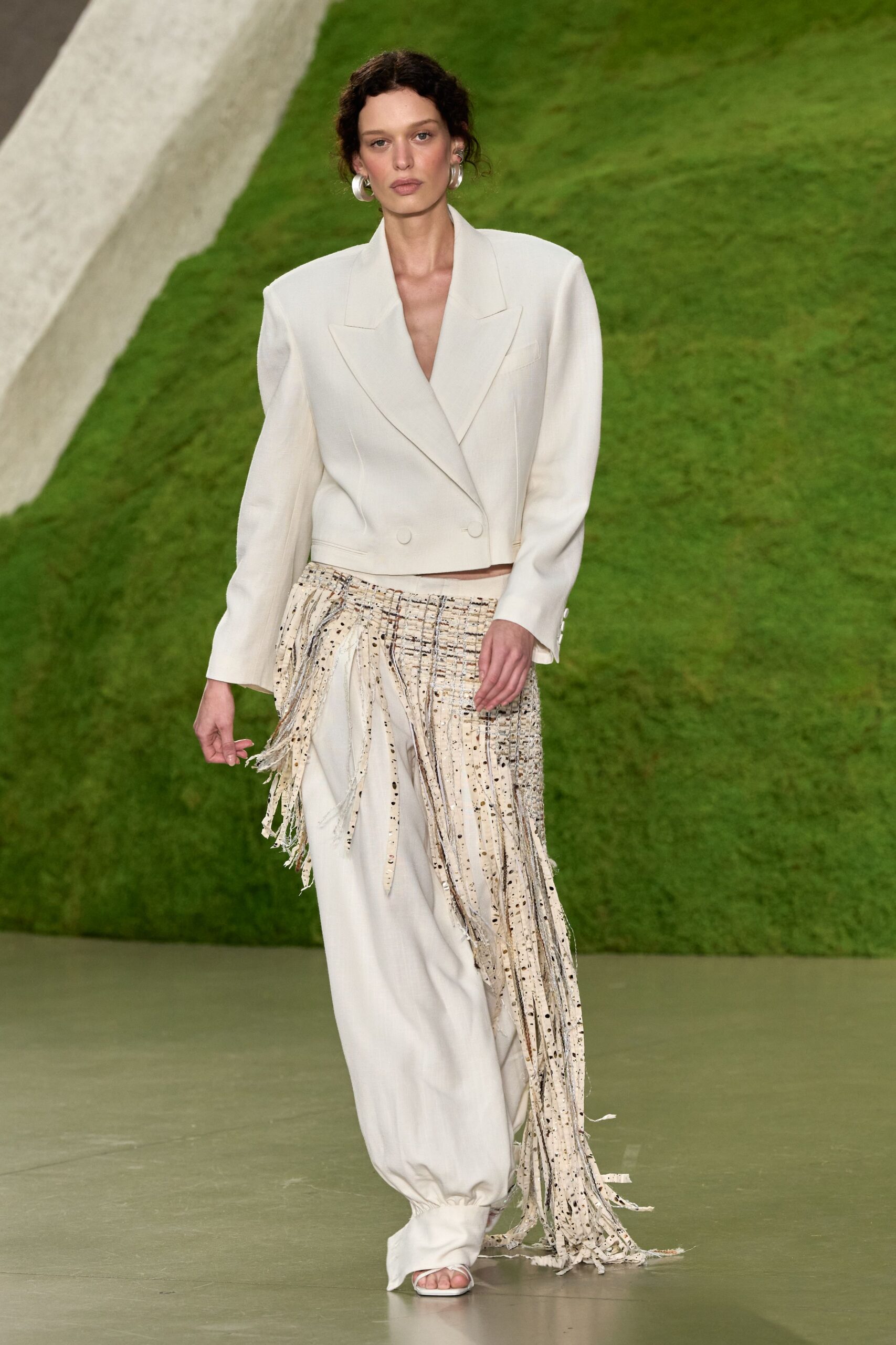 Ivory cropped double-breasted blazer with peaked lapels and padded shoulders. Large silver hoops. Ivory wide-leg trousers with a swaying panel of speckled tweed fringe. White square-toe sandals peeking under cuffed hems. Crossing a sculpture garden lawn before the speeches start.