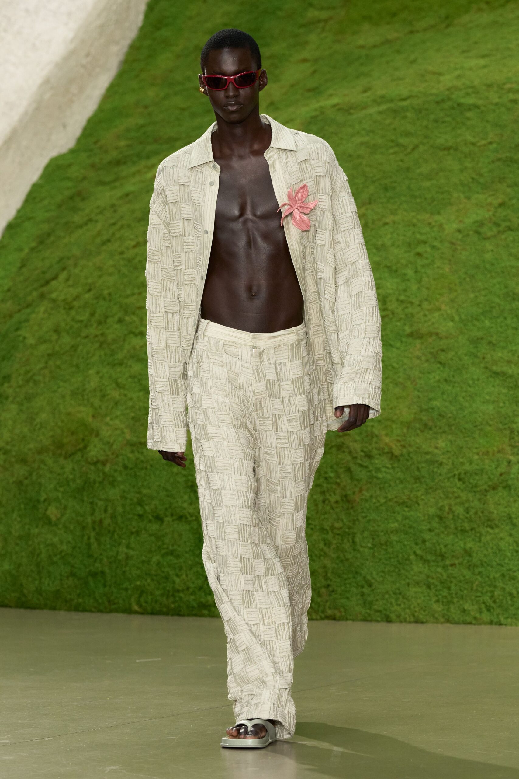 Narrow red wraparound sunglasses with pointed outer corners. Open cream shirt in raised basket-weave, a pink flower pinned at the chest. Matching loose trousers, the woven blocks running uninterrupted to the hem. Dark thong sandals on thick square soles. Walking across a clipped lawn after the wedding tents came down.