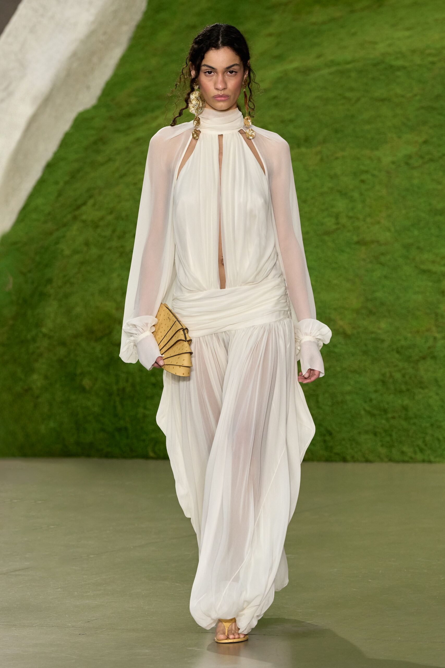 Ivory chiffon gown with a high wrapped neck and plunging slit. Sheer cape sleeves ending in gathered cuffs. Oversized gold coin drops brushing the jaw. Fan-shaped tan clutch, pinholed like ostrich leather. Flat gold thong sandals peeking from the hem. Walking across a hotel lawn after midnight, shoes in the wrong register.