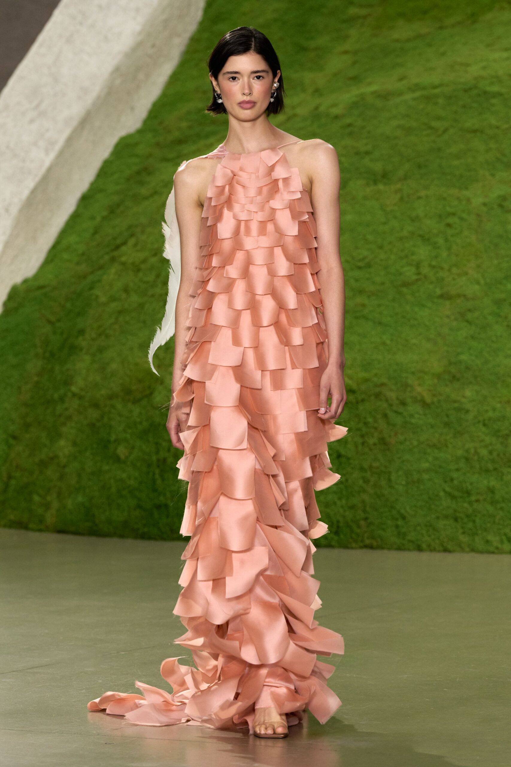 Blush satin column gown layered all over with staggered square flounces. Single white feather wing trailing from one shoulder blade. Barely there nude sandals, one toe peeking under the hem. Small silver drop earrings. Crossing a botanical atrium after the string quartet has packed up.