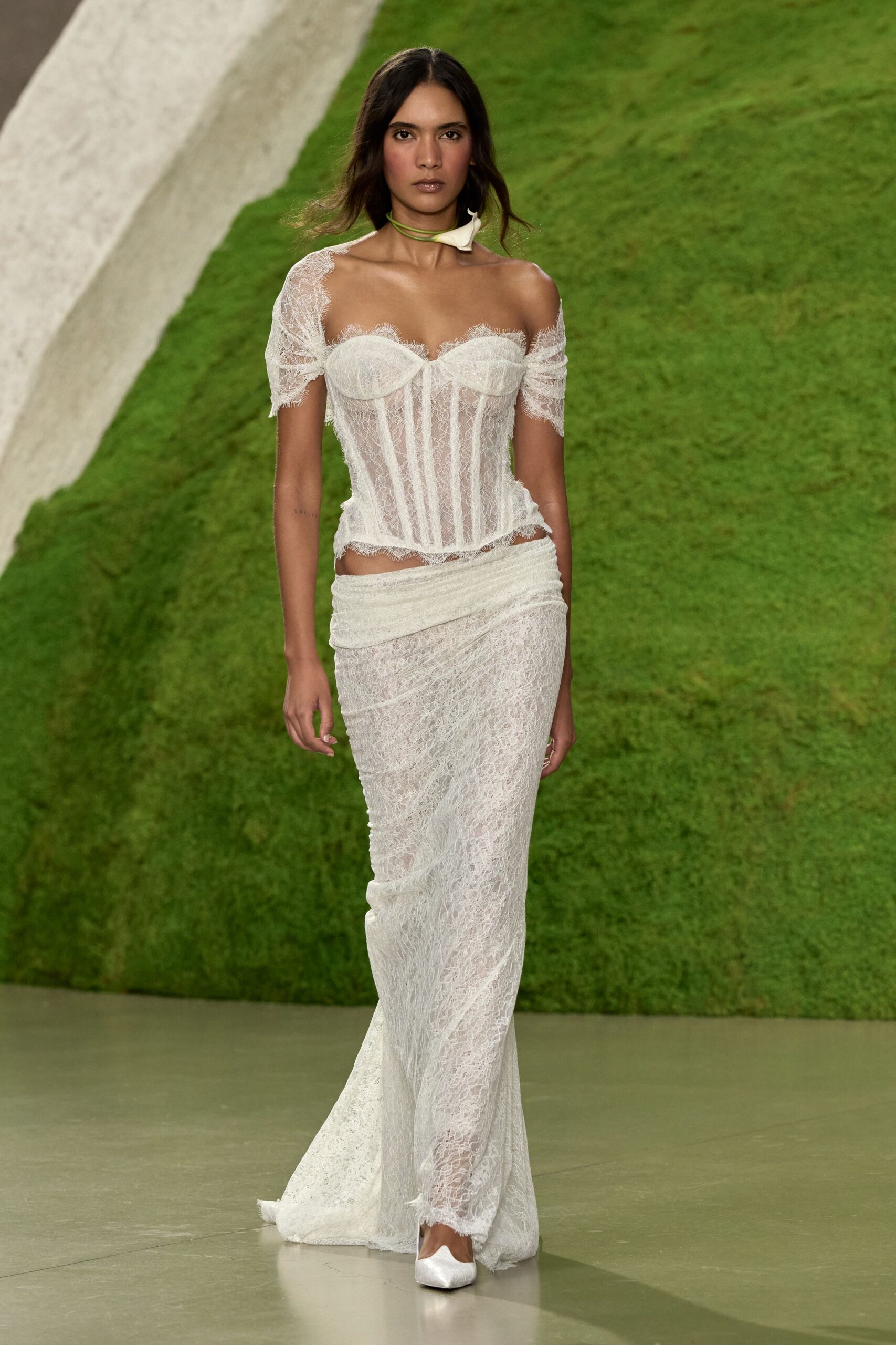 Green cord choker tied with a small white calla lily. Ivory lace corset top, sheer boned panels and eyelash cups slipping off-shoulder. Matching lace column skirt, wrapped low with a folded waistband. White satin flats with square toes. For crossing a lawn after the ceremony, heels already in hand.