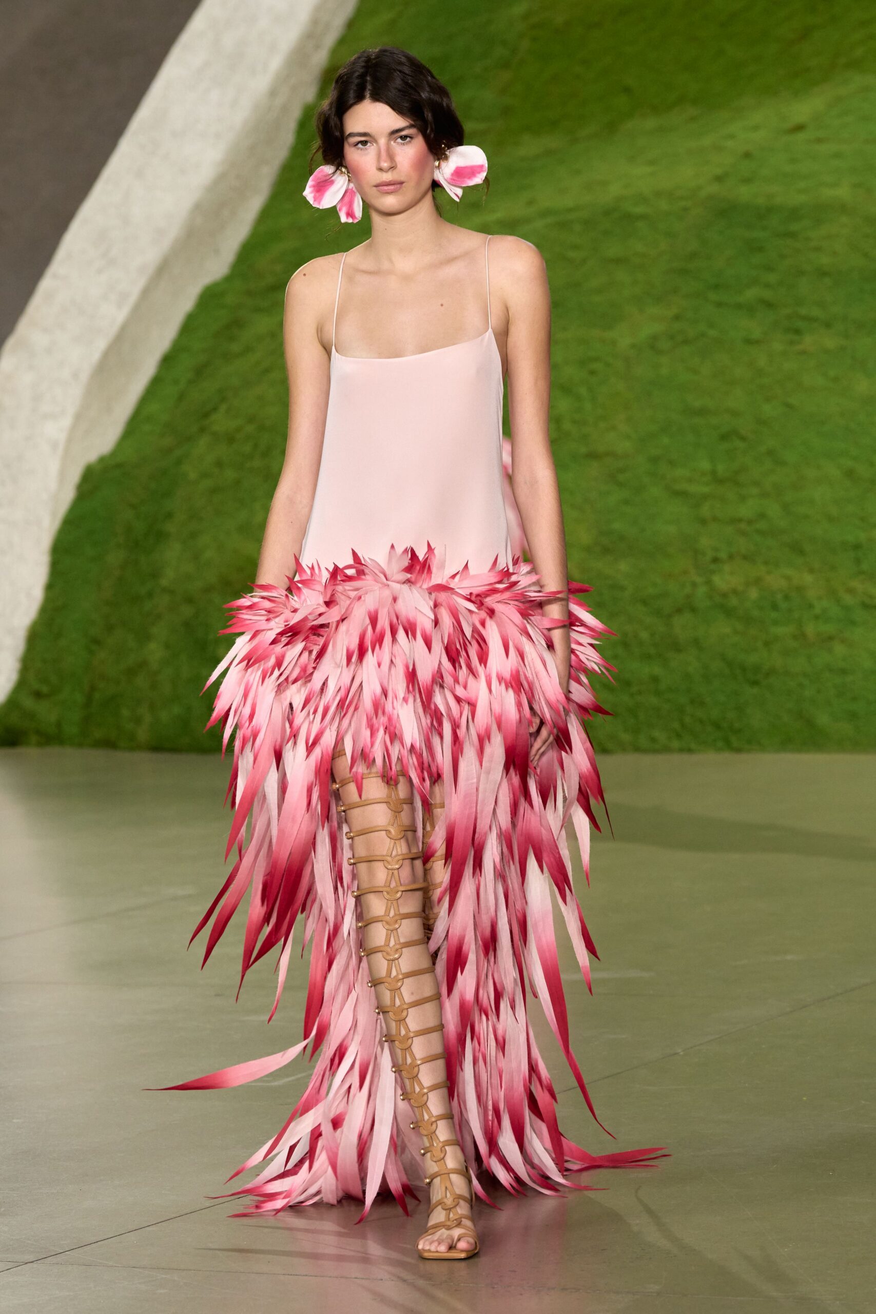 Oversized pink-and-white petals cupping both ears. Blush slip dress on spaghetti straps, exploding into dip-dyed feather fringe. Tan gladiator sandals laced to the knee with ringed cords. Crossing a lawn after the wedding band has packed up.