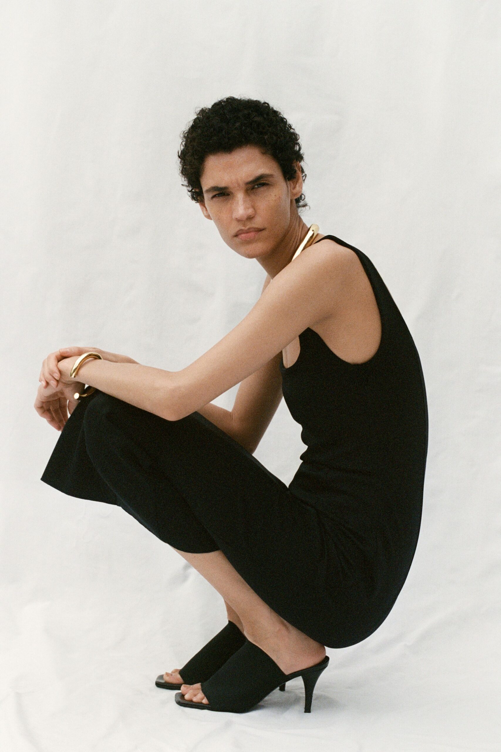 Black jersey jumpsuit with a low scooped armhole and cropped wide legs. Thick gold collar curving off the shoulder. Matching gold cuff, smooth and weighty. Black stretch mules on a narrow kitten heel. Morning after a hotel wedding, still barefoot in your head.