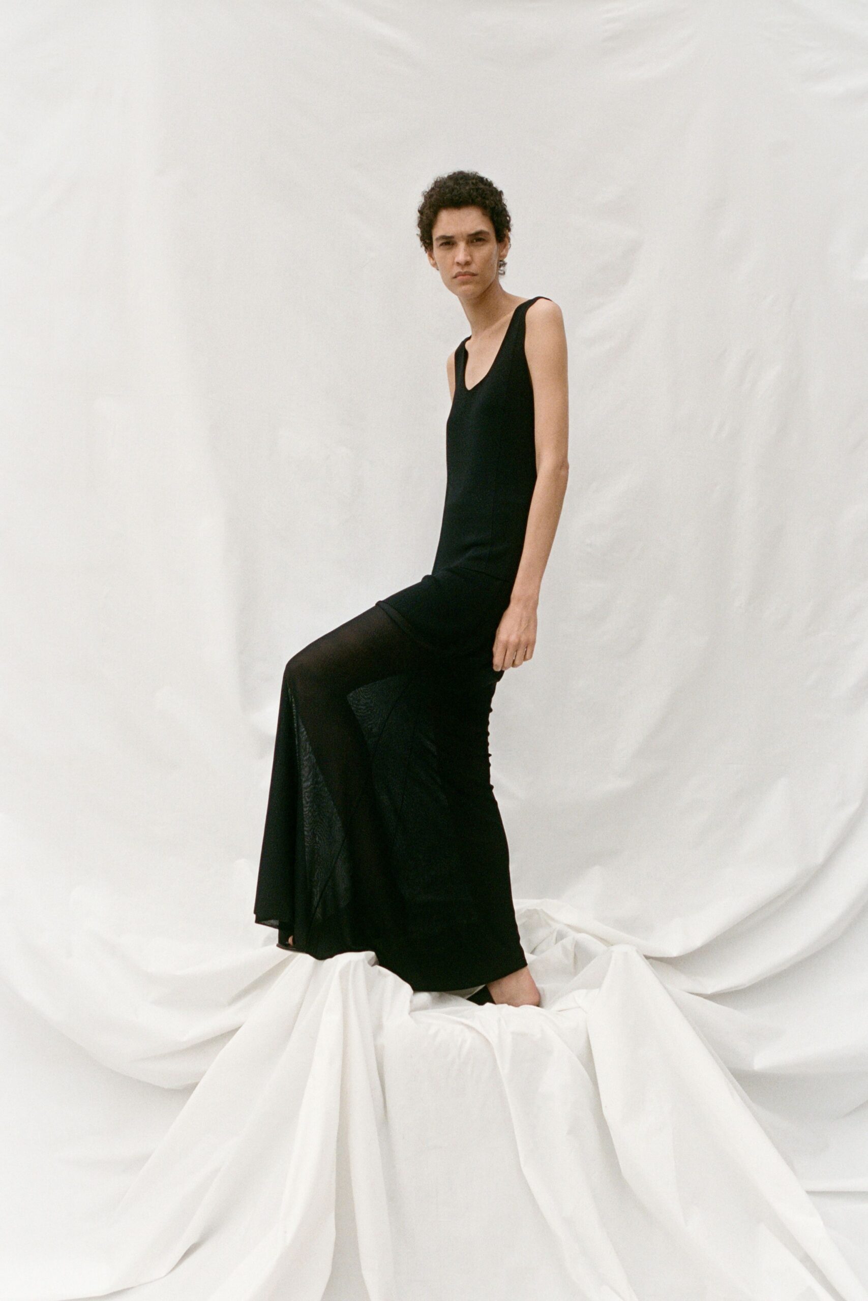 Black column dress in matte jersey, scooped low and slit to mid-thigh. Black pointed toes barely showing under the pooled hem. Curtains pinned up for painting, one window cracked against the heat.