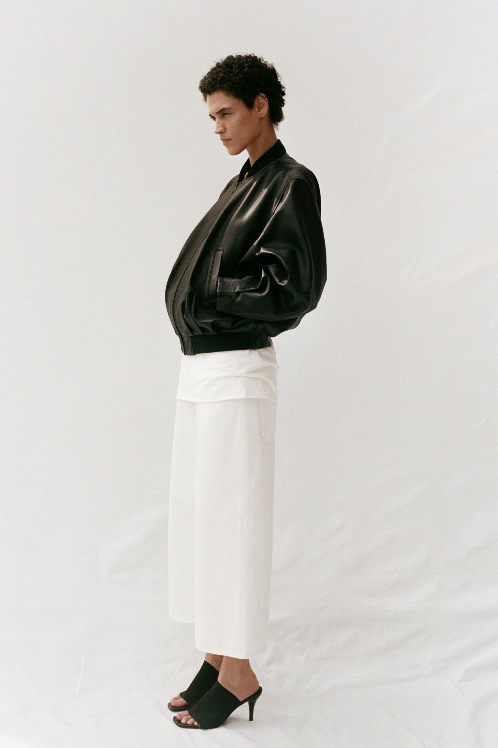 Black leather bomber, balloon sleeves collapsing into a ribbed hem. White underlayer hanging below the jacket in a clean band. Ivory wide-leg culottes with a soft vertical rib. Black ankle socks. Black peep-toe mules on a narrow kitten heel. First ferry out, carrying coffee carefully across the wet deck.