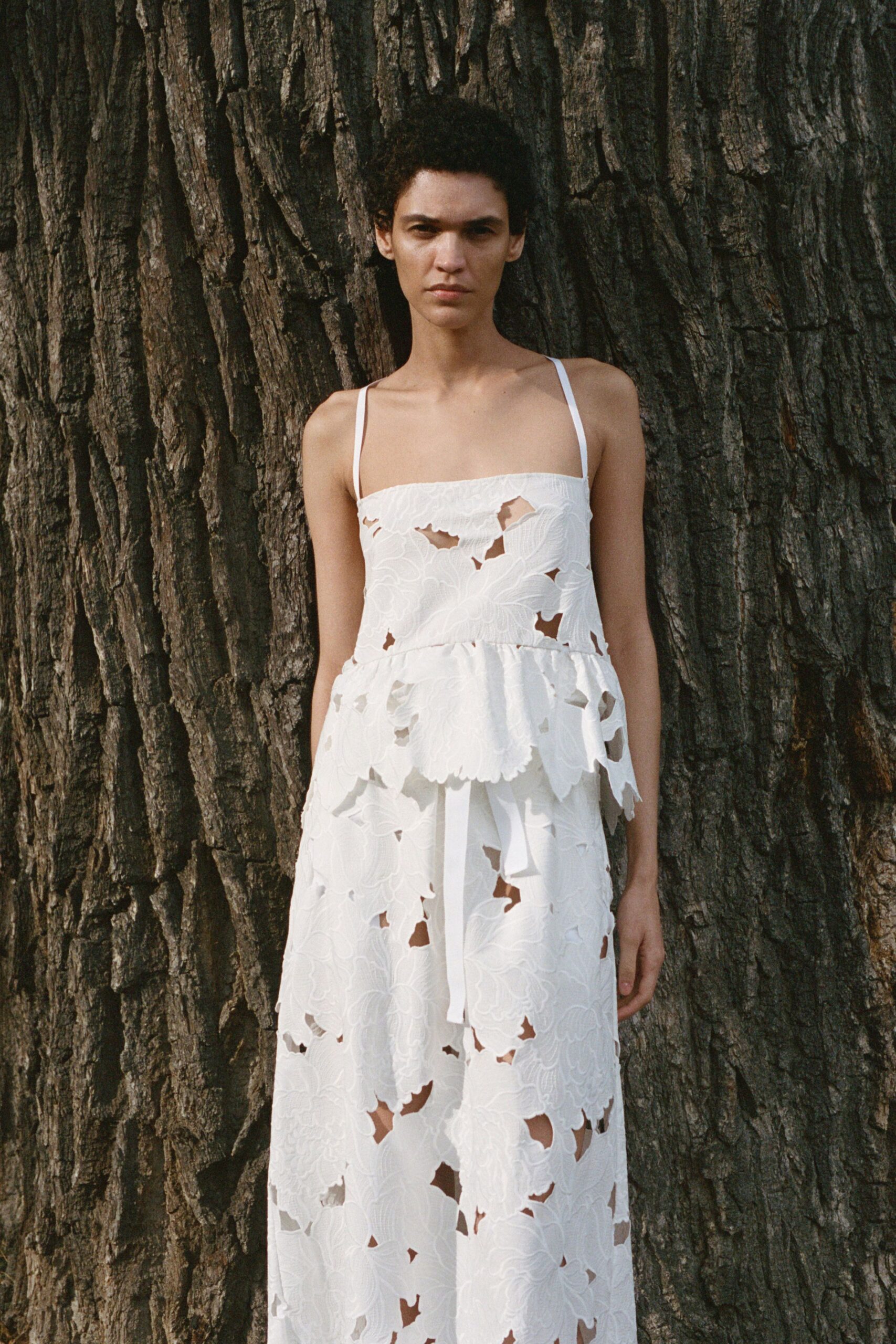 White spaghetti-strap dress in floral cutwork, peplum waist and long ribbon tie. Standing under a park tree after the grass has gone straw-dry.
