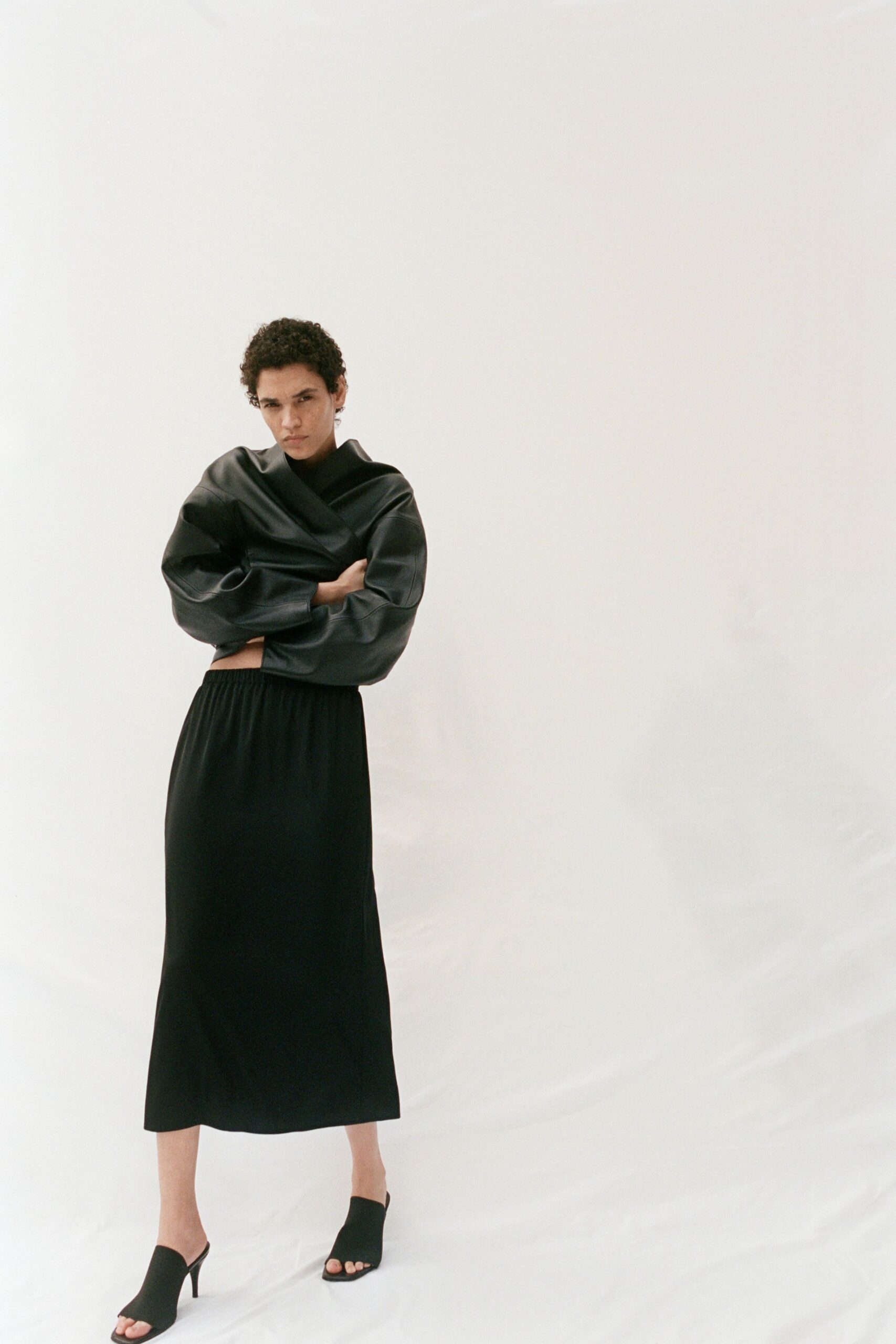 Black leather wrap top with sculpted sleeves and a cropped raw edge. Black column skirt in fluid crepe, gathered softly at the waist. Black peep-toe mules on slim stiletto heels. First meeting in an empty loft before the furniture arrives.