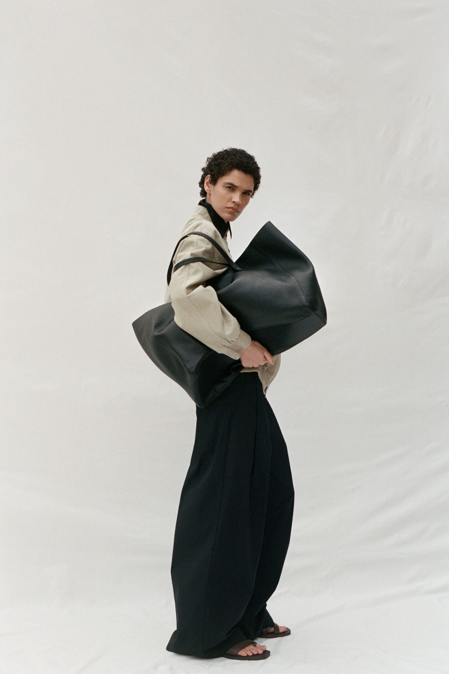 Bone cotton blouson with gathered cuffs and black binding tracing the shoulder. Black knit turtleneck tight to the jaw. Oversized folded black leather bag tucked under both arms. Black wide-leg trousers breaking into a soft puddle at the hem. Flat black thong sandals. Carrying too much through an airport corridor before sunrise.