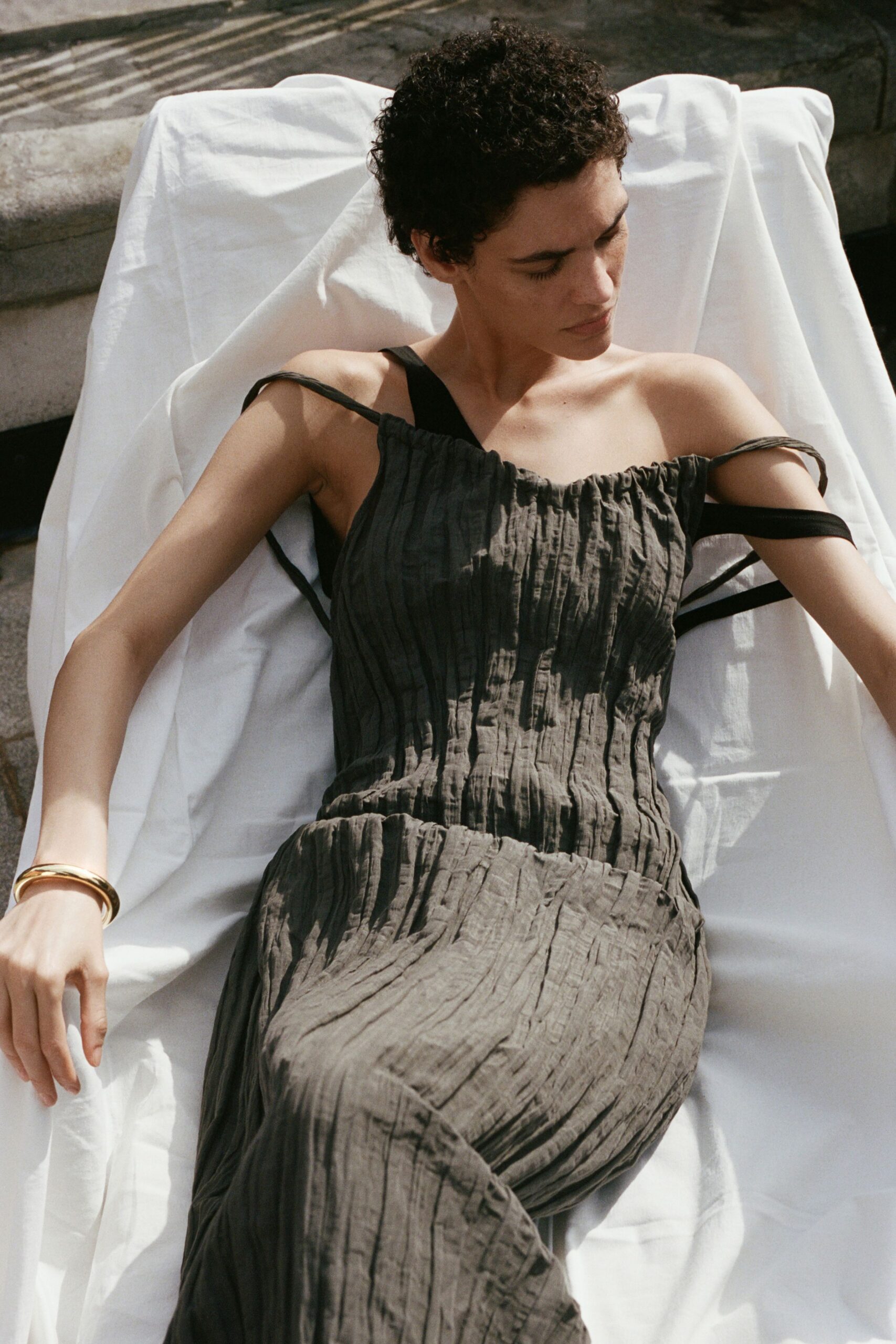 Charcoal crinkled dress with a ruched neckline and dropped shoulder straps. Black one-shoulder underlayer cutting a clean band across the upper arm. Single gold bangle, smooth and weighty. Sun on the terrace lounger after everyone else has gone inside.