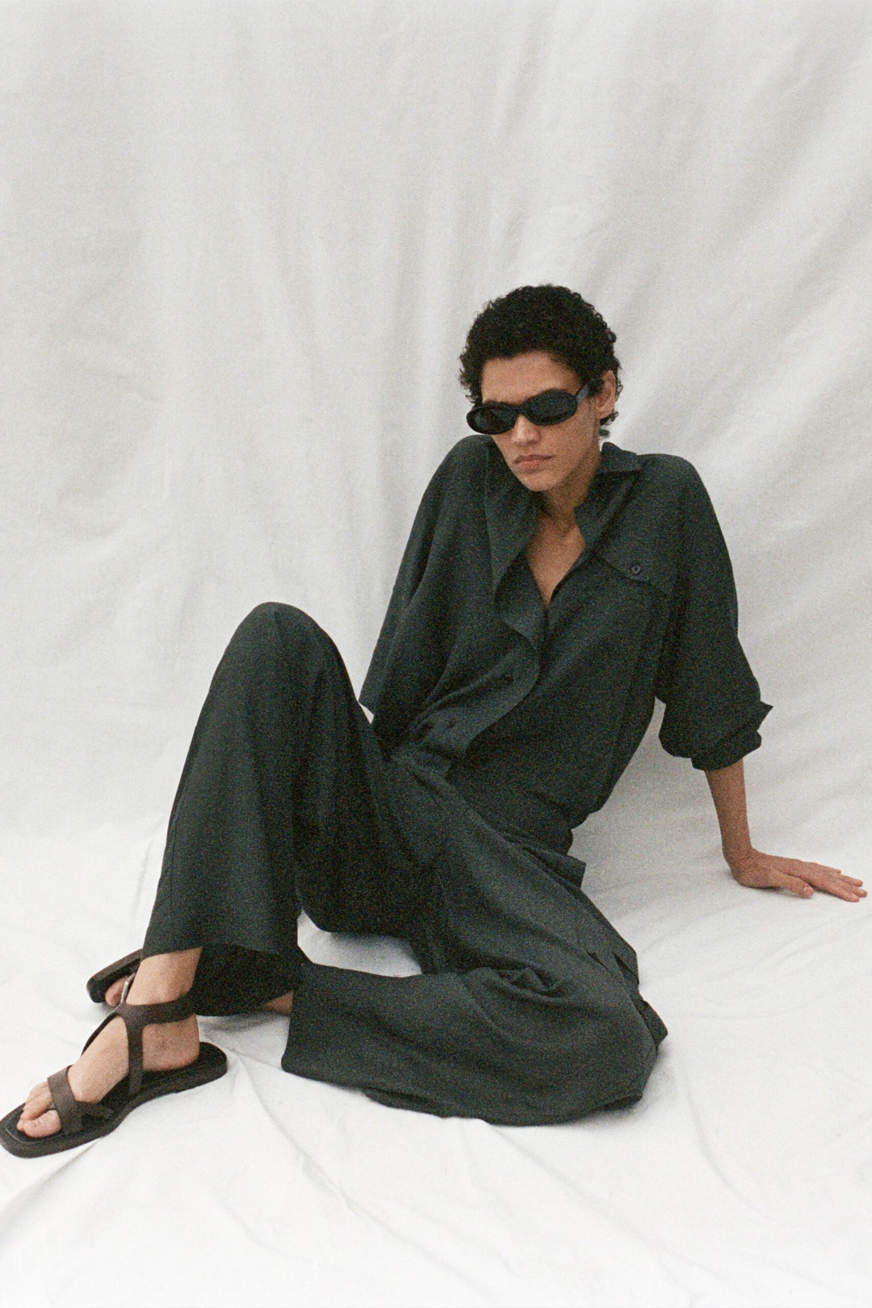 Narrow black oval frames hugging close to the face. Charcoal fluid button-up shirt, one flap pocket fixed with a small rivet. Matching wide-leg trousers in the same draped weave. Flat black T-strap sandals with thick foam soles. Sunday heat, blinds half shut, nowhere you need to answer to.