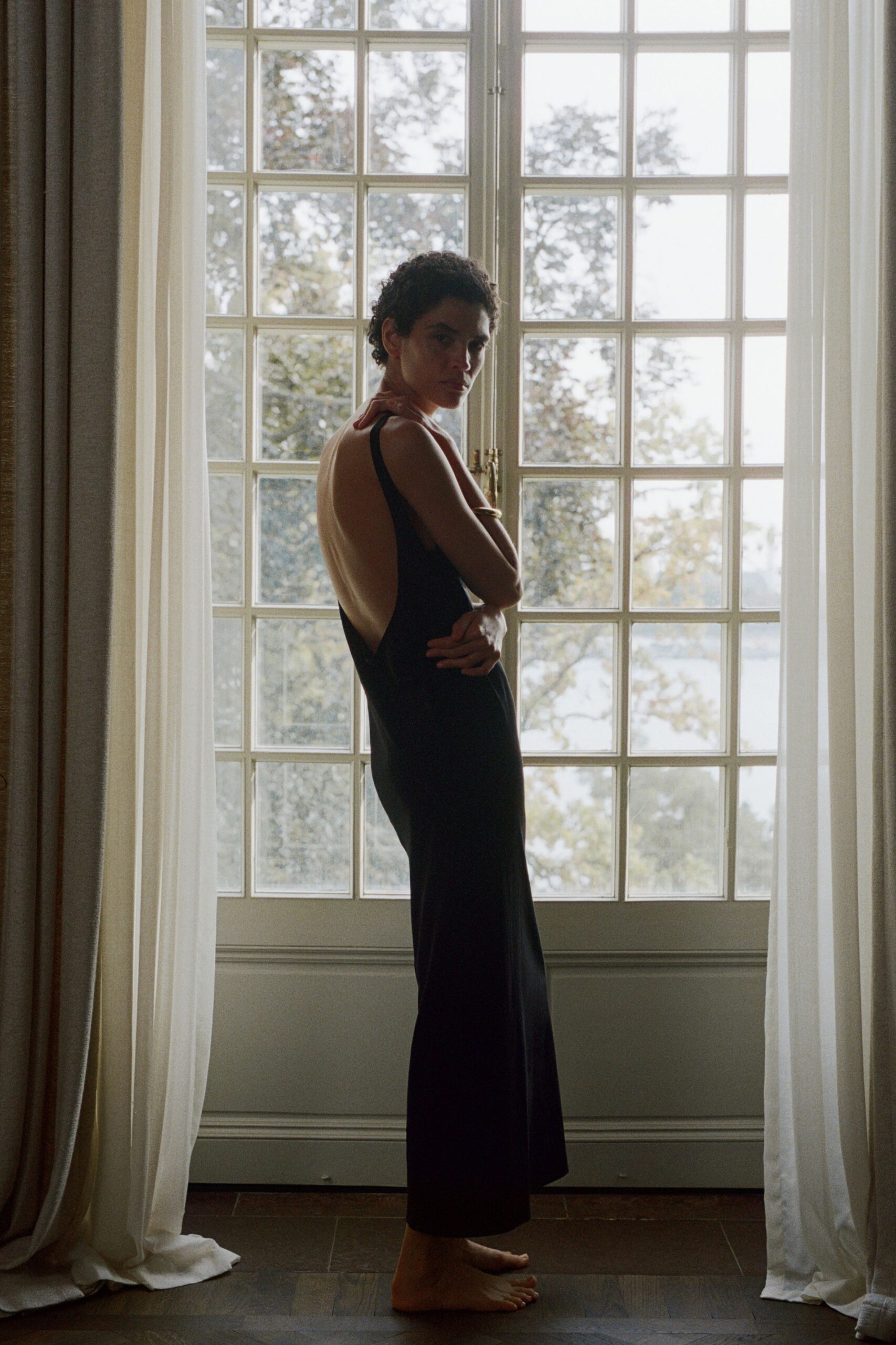 Black bias-cut slip dress, sides scooped nearly to the waist at the back. Slim gold bangle catching the window light. Pausing at a lakehouse window before anyone else wakes.