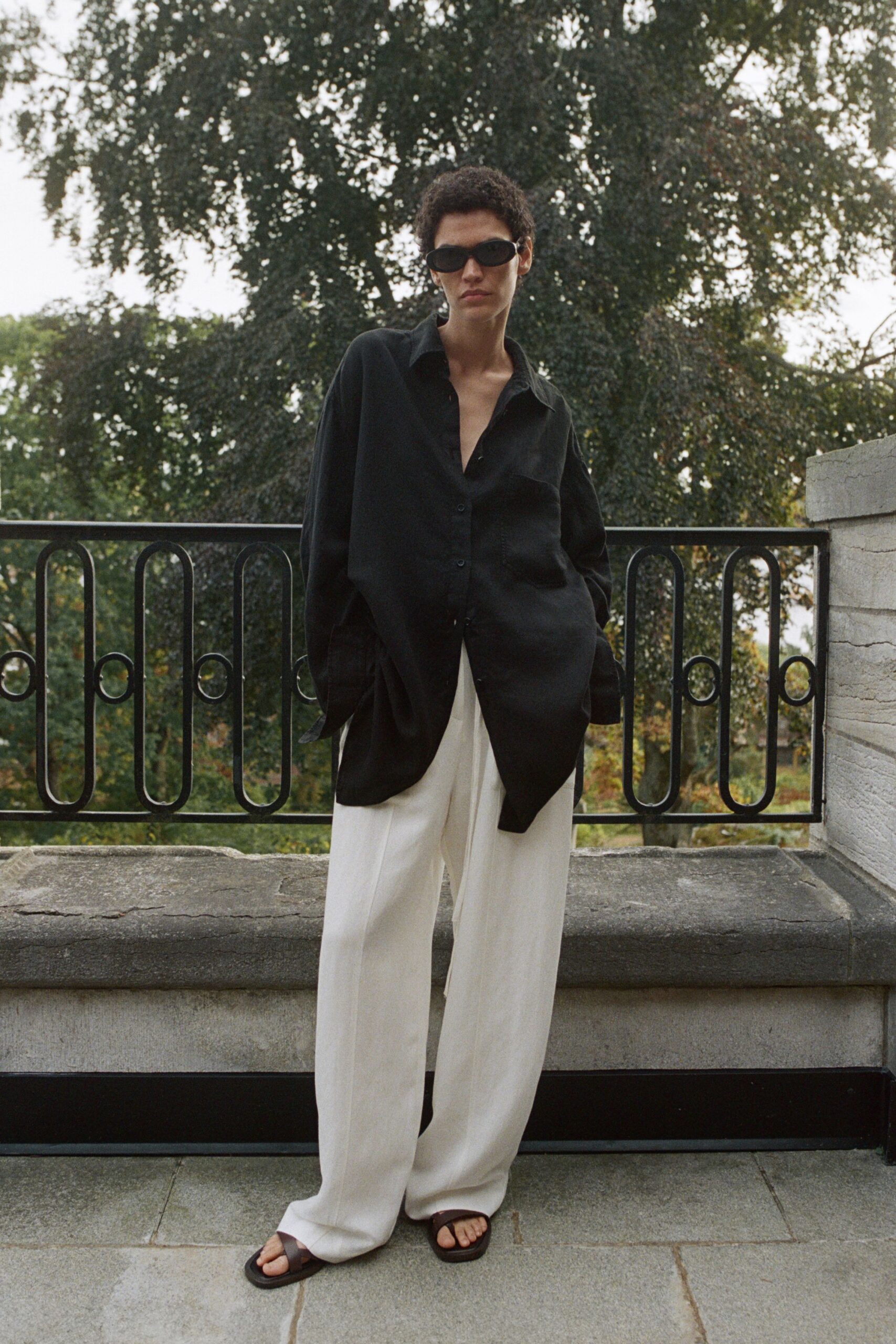Narrow black wraparound sunglasses. Oversized black shirt in fluid matte silk, one chest pocket sagging slightly. Chalk wide-leg trousers with a pressed crease and pooling hem. Dark brown leather slide sandals, square-toed. Stepping onto a hotel terrace before the afternoon storm breaks.