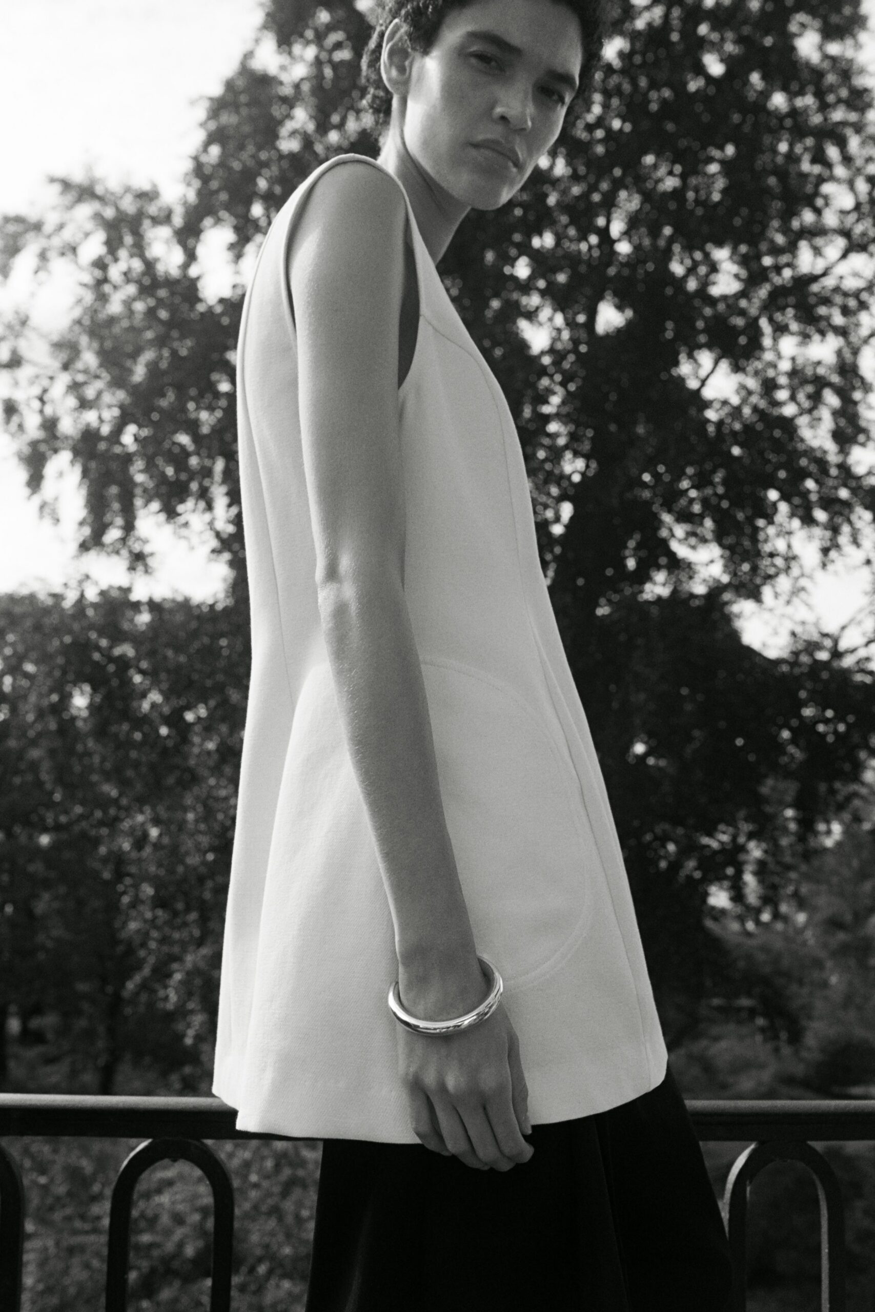 White structured tunic, deep armholes and curved welt pockets at the hip. Black skirt falling straight beneath the long tunic hem. Thick silver bangle with a softly dented surface. Leaning over a park railing after a wedding has thinned out.