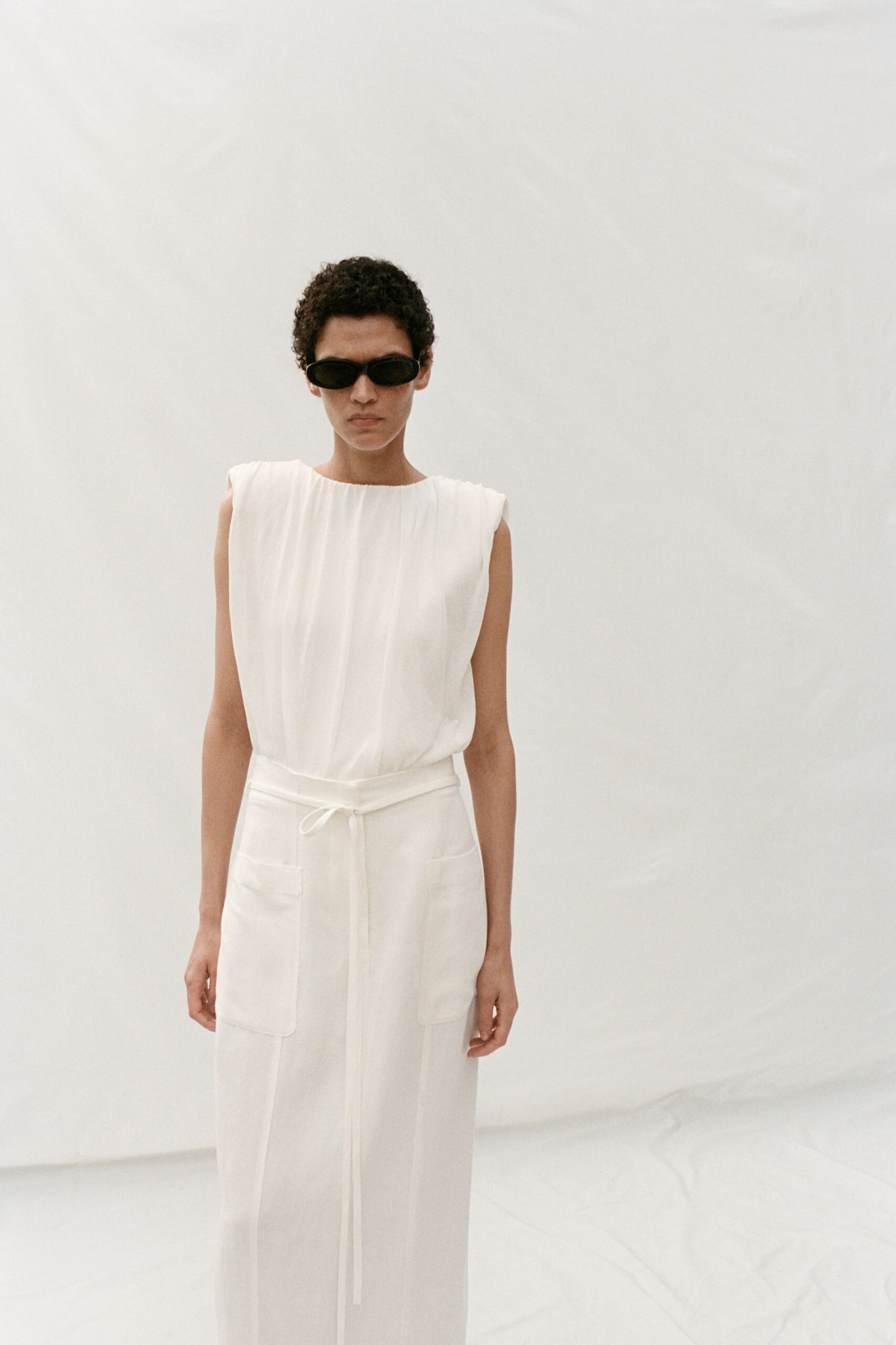 Narrow black wrap sunglasses. Ivory sleeveless column dress with softly gathered bodice and tied sash. Oversized patch pockets set low on the skirt front. Morning checkout, hotel robe mood but nothing actually soft.