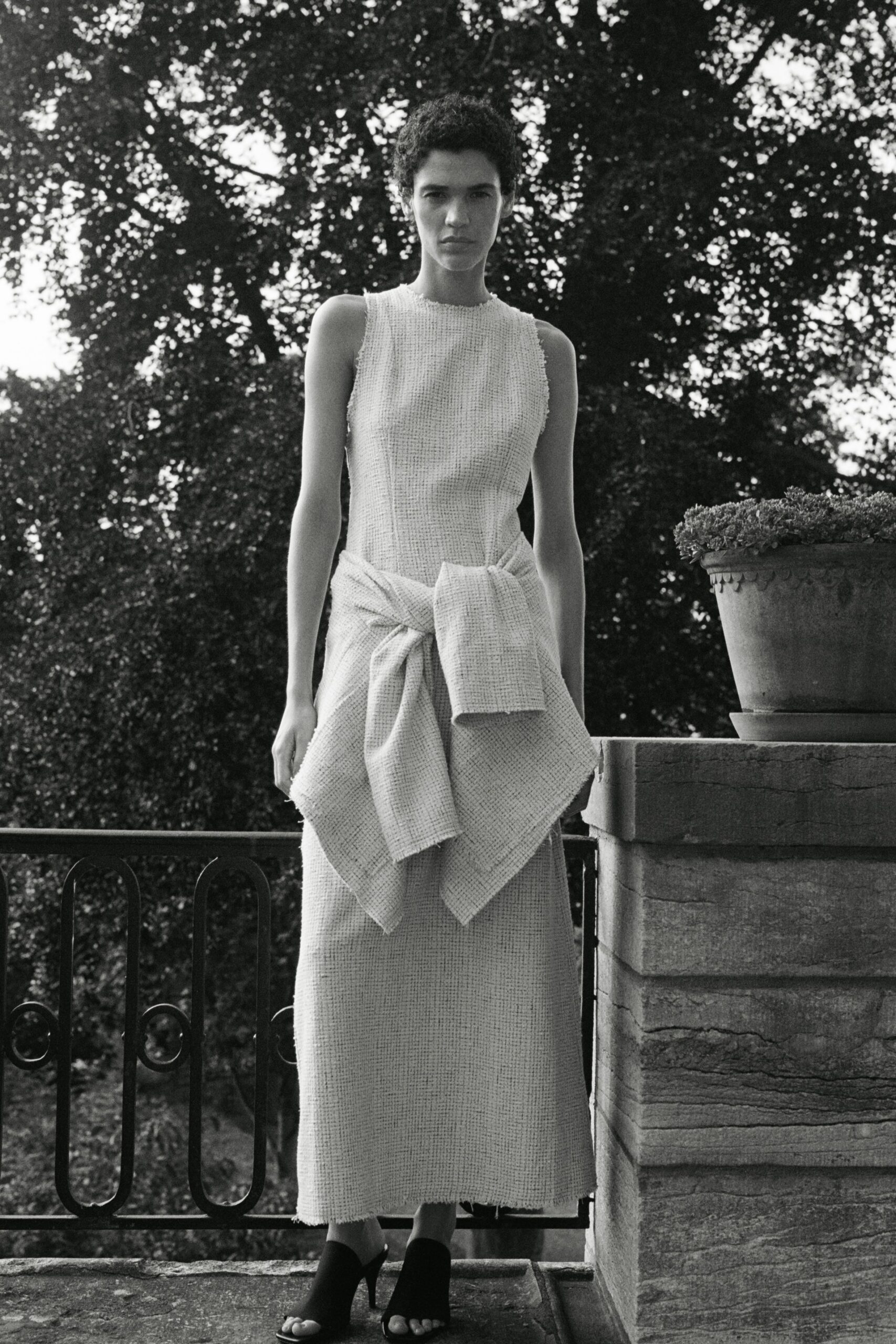 Sleeveless cream basket-weave column dress with a raw, nubby edge. Matching knit tied low at the waist, one panel hanging longer. Black peep-toe mules with a narrow curved heel. On a stone terrace before dinner, the garden already going dark.