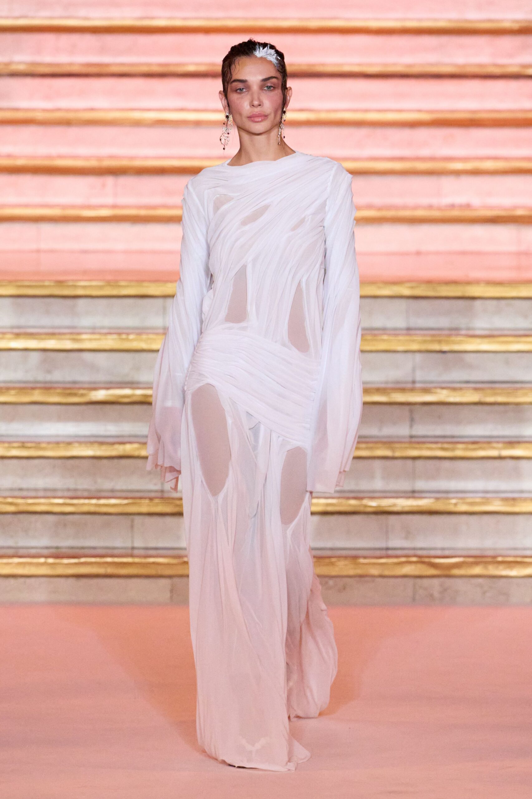 Blush chiffon gown in shredded, spiraling bands over sheer cutout panels. Long crystal drop earrings brushing the jaw. Descending a painted staircase after the wedding guests have gone.