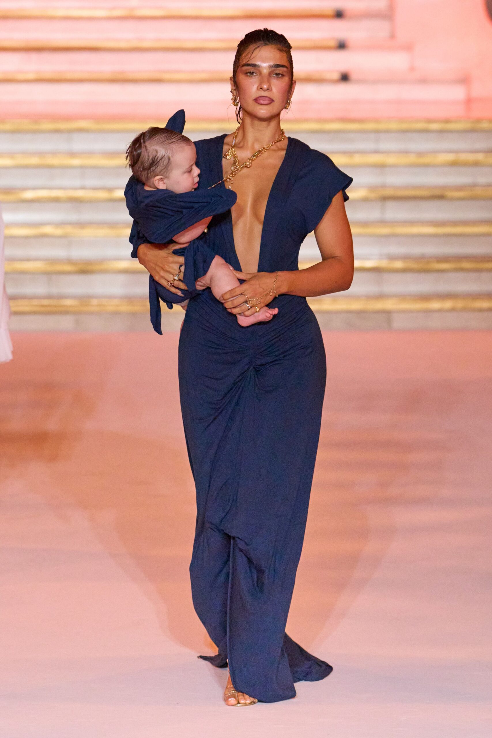 Ink-blue jersey gown with a plunging V and ruched front knot. Gold barely-there sandals. Heavy twisted gold chain crossing the collarbone. Fine gold chains at the wrist. Matching navy sling carrying a baby at the hip. Walking into a wedding reception after the babysitter canceled.