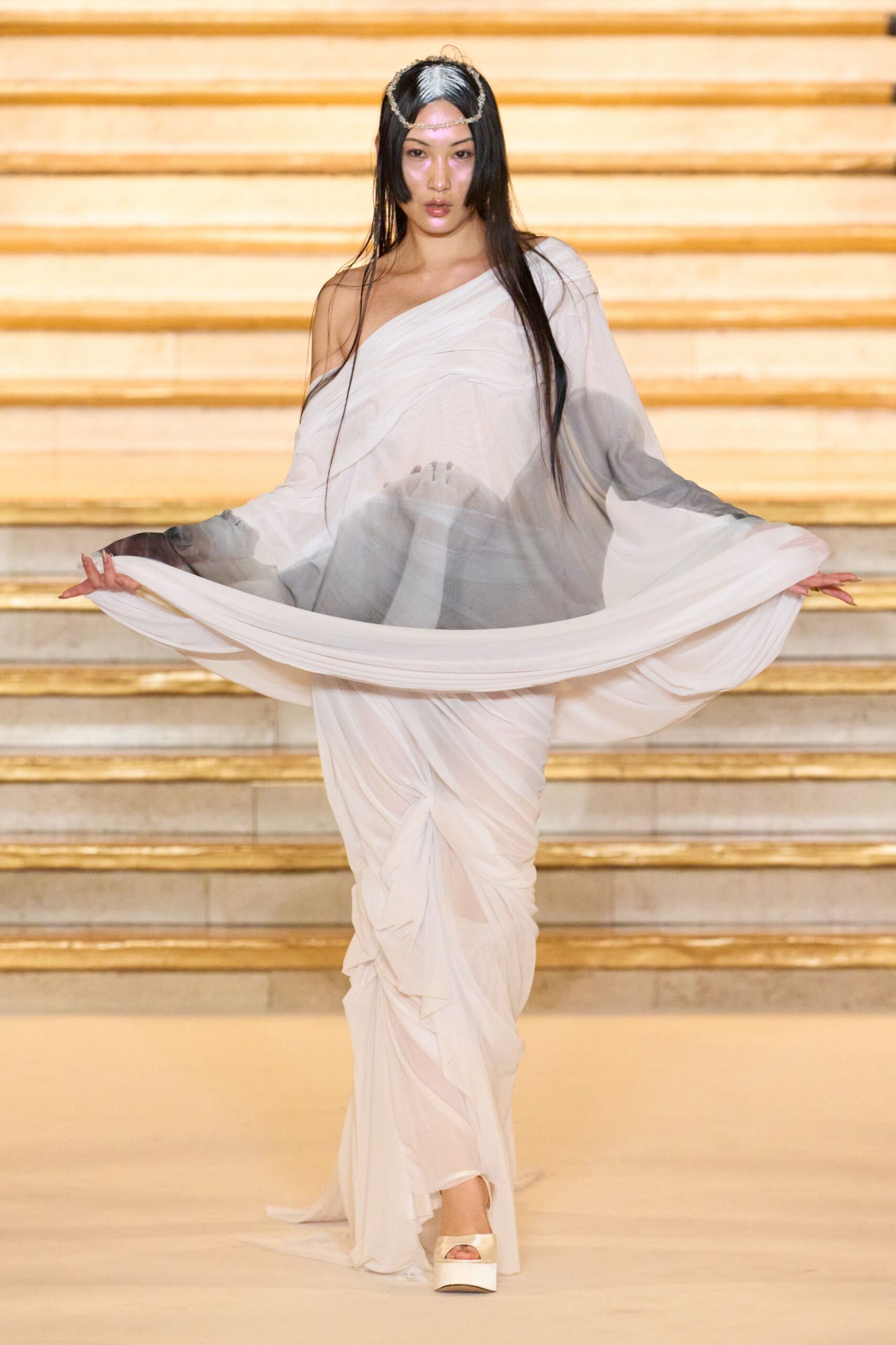 Fine gold chain dipping across the forehead and over the crown. Ivory chiffon gown, one-shoulder and densely ruched into hanging folds. Cream satin platforms with a blunt peep toe. Crossing a hotel staircase after the ceremony, still holding the train wide.