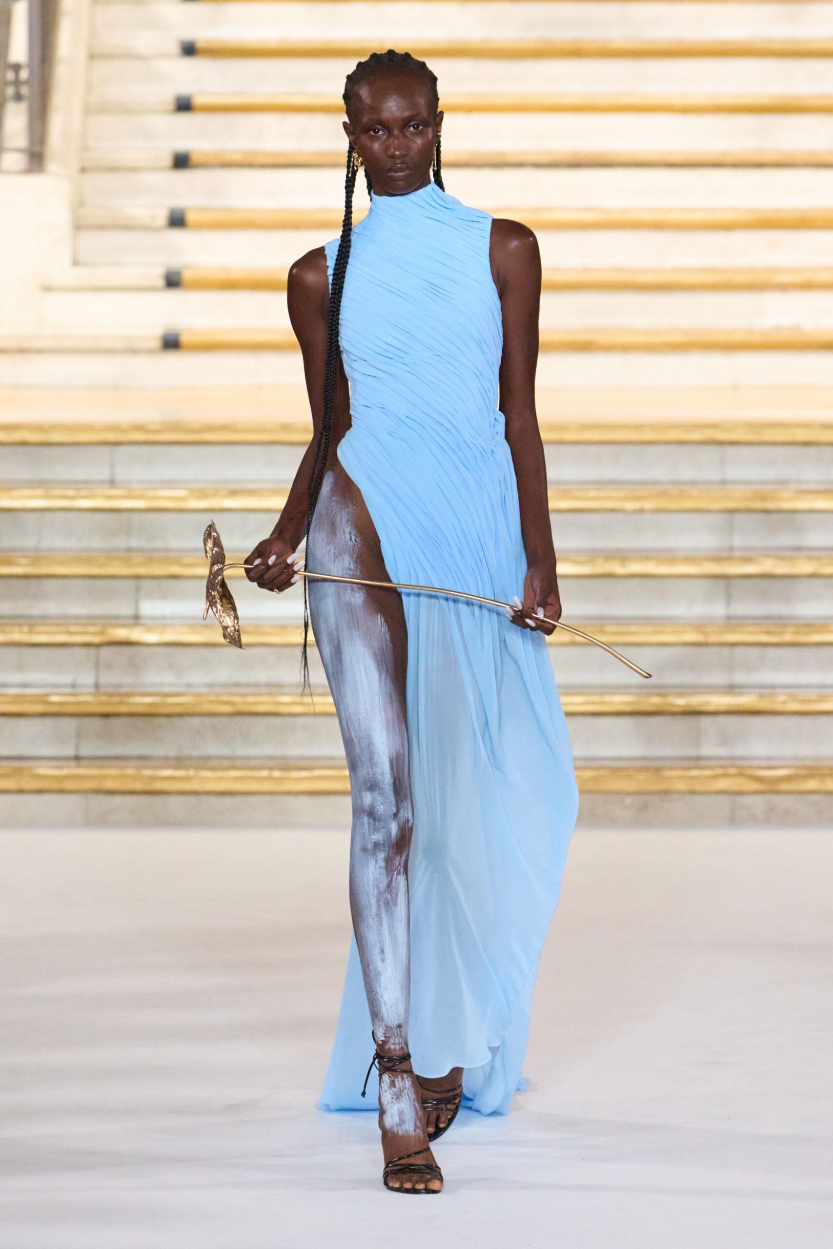 Sky-blue pleated jersey gown, one side carved to the hip. White brushstrokes dragged down one bare leg. Black lace-up sandals with thin cords crossing the ankle. Long-stemmed metal flower held like a wand. Descending a marble staircase after midnight with pollen nowhere in sight.