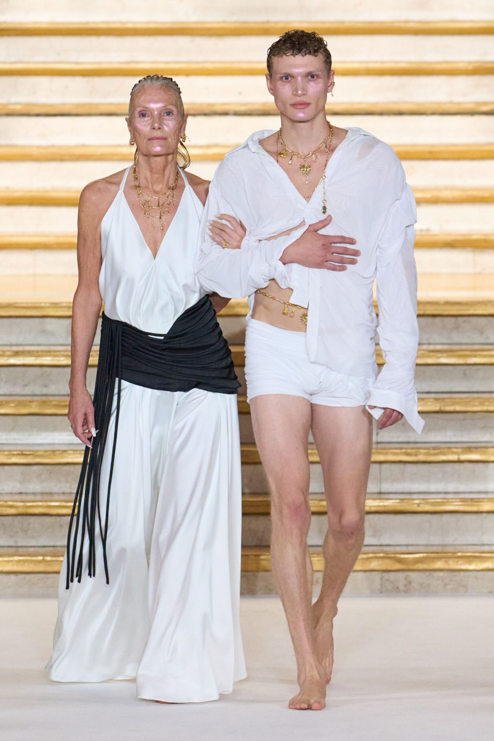 White gauze blouse knotted off-center, one side cut open at the waist. White ruched shorts sitting low on the hipbones. Gold charms draped at the neck and bare midriff. Unshod feet, toes fully exposed. Walking down hotel stairs after a dawn swim, shirt still salt-soft.