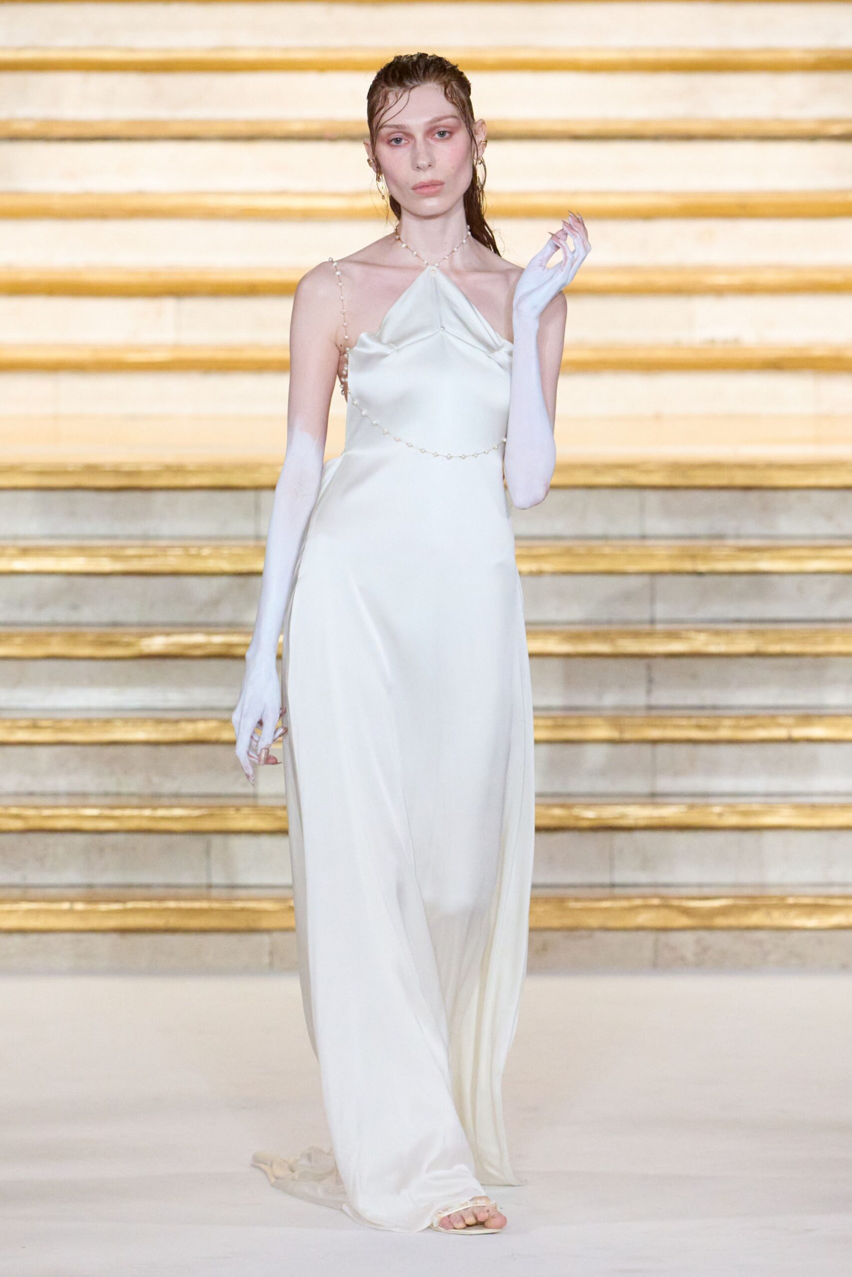 Ivory satin column gown with a folded halter point and low train. Pearl-beaded straps and waist chain, one strand dipping off-center. White opera gloves fading away around the upper arm. Ivory slide sandals peeking from the hem. For crossing a marble lobby after the ceremony, hair still wet.