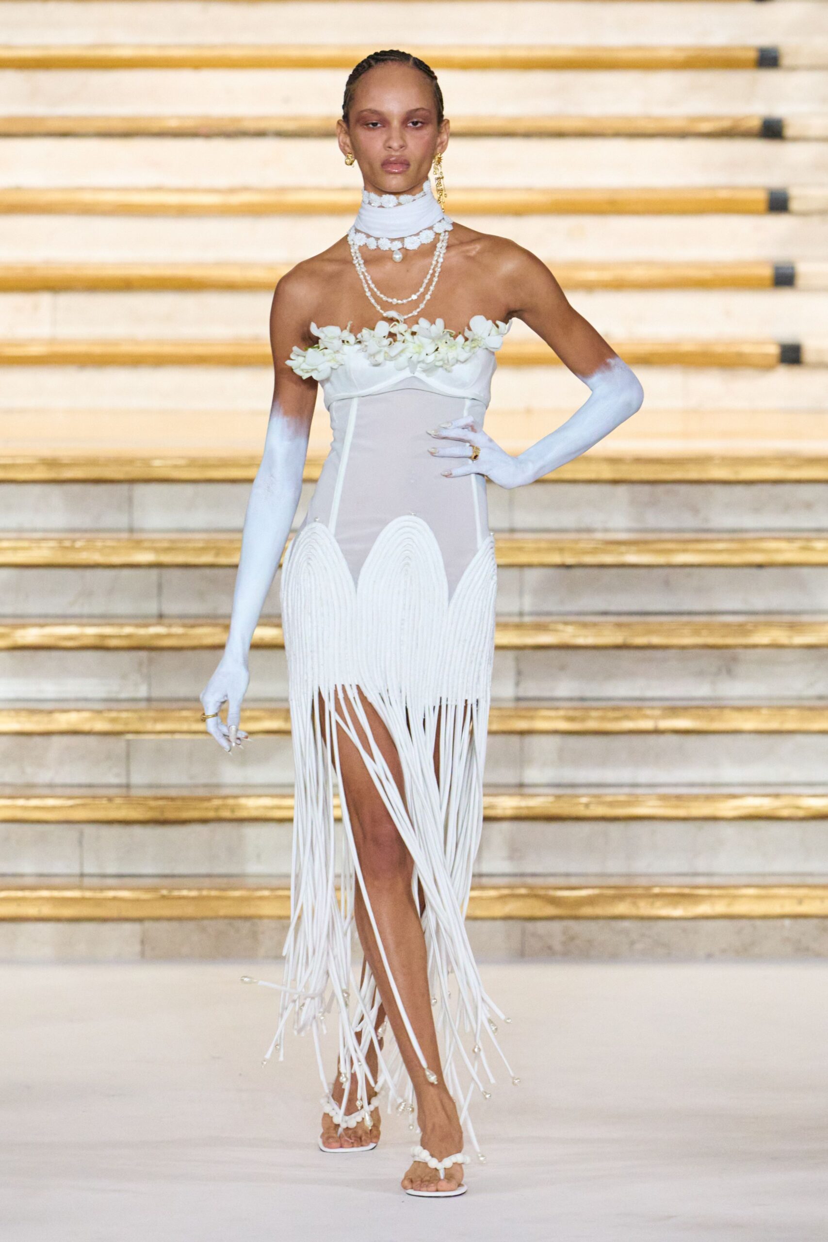 Pale blue neck wrap edged in tiny white blossoms. Sheer white column dress, orchid-trimmed bust and long cord fringe. Powder-blue gloves fading to chalk at the upper arm. White thong sandals with petal clusters at the straps. Walking down hotel stairs after a beach wedding that ran past sunset.