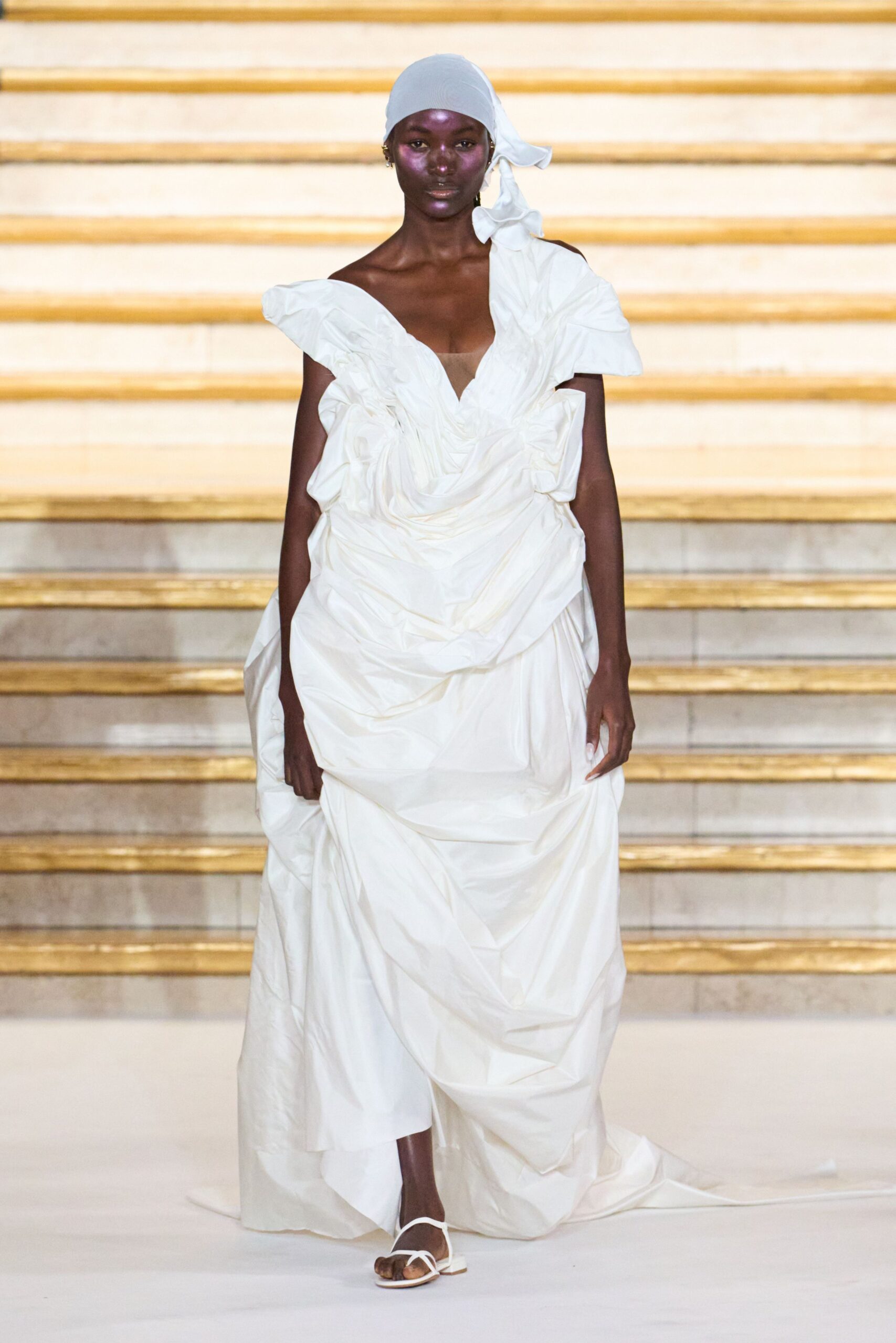 Pale dove headscarf tied low, one long tail knotted at the side. Ivory taffeta gown, densely ruched into swollen folds and an off-shoulder plunge. White thong sandals with flat square soles. City hall steps after the ceremony, bouquet paper going soft in your hand.