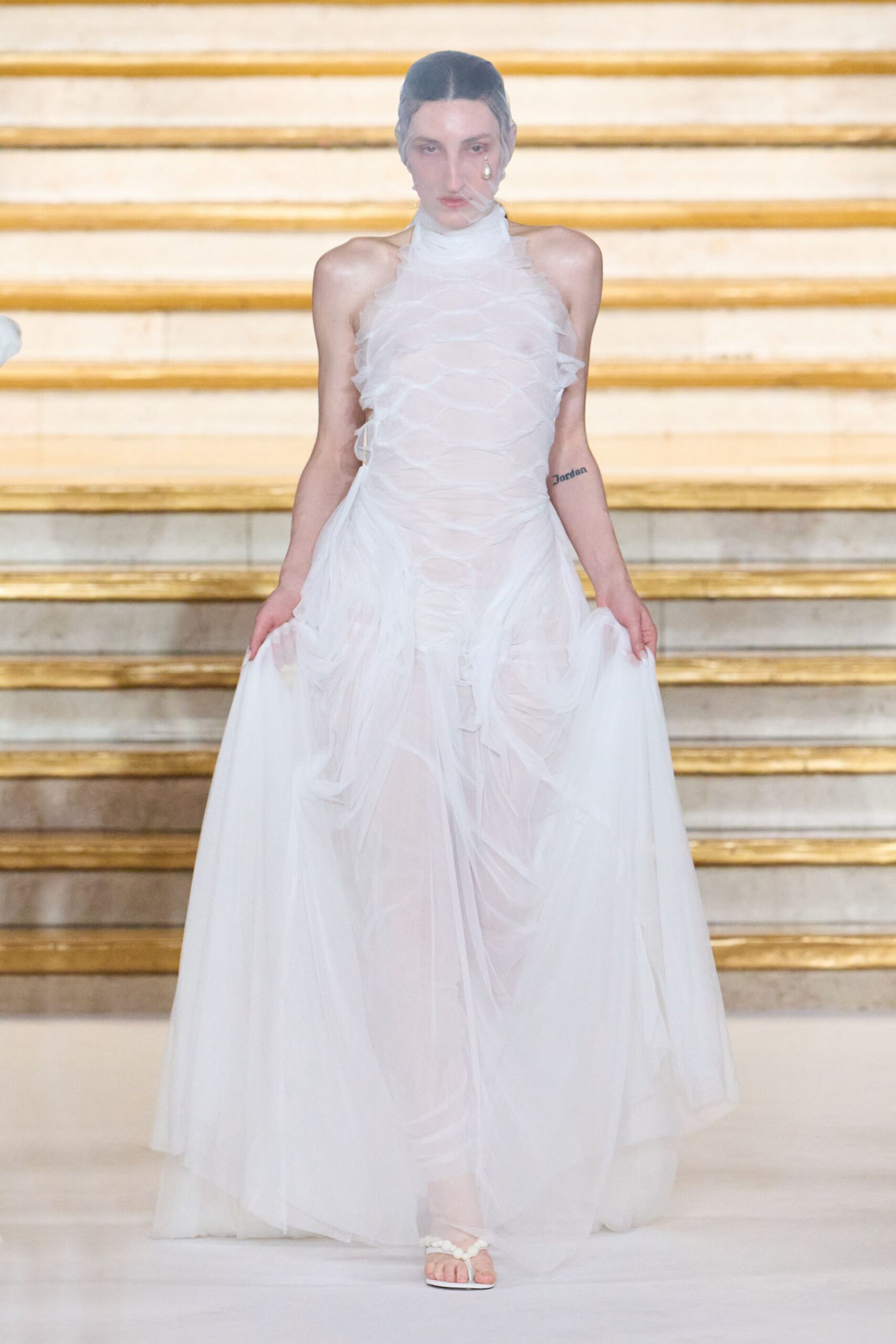 Sheer face veil pinned close, one pearl drop caught at the cheek. White halterneck tulle gown, ruched through the torso with fluttering ruffle edges. White thong sandals with clustered bead straps. Crossing a marble staircase after the ceremony, still holding up the hem.