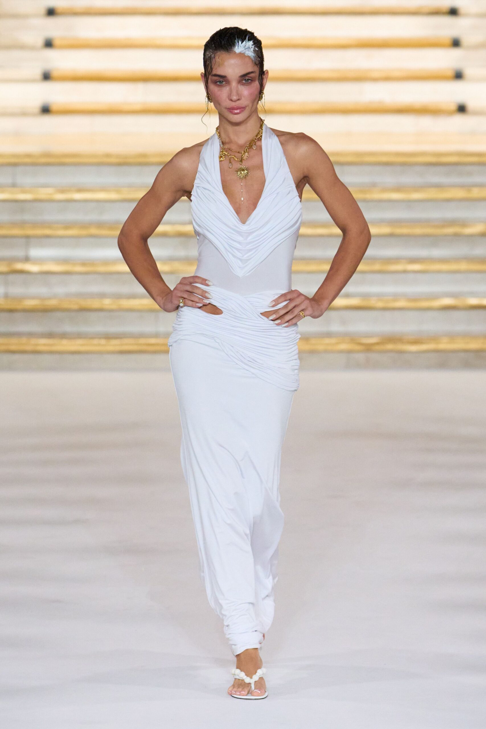 Layered gold charms with a pearl drop hanging to the sternum. White jersey halter gown, ruched cowl bodice and twisted hip cutout. White toe-ring sandals with a knotted bead strap. Walking down hotel steps after a saltwater wedding, hair still wet.