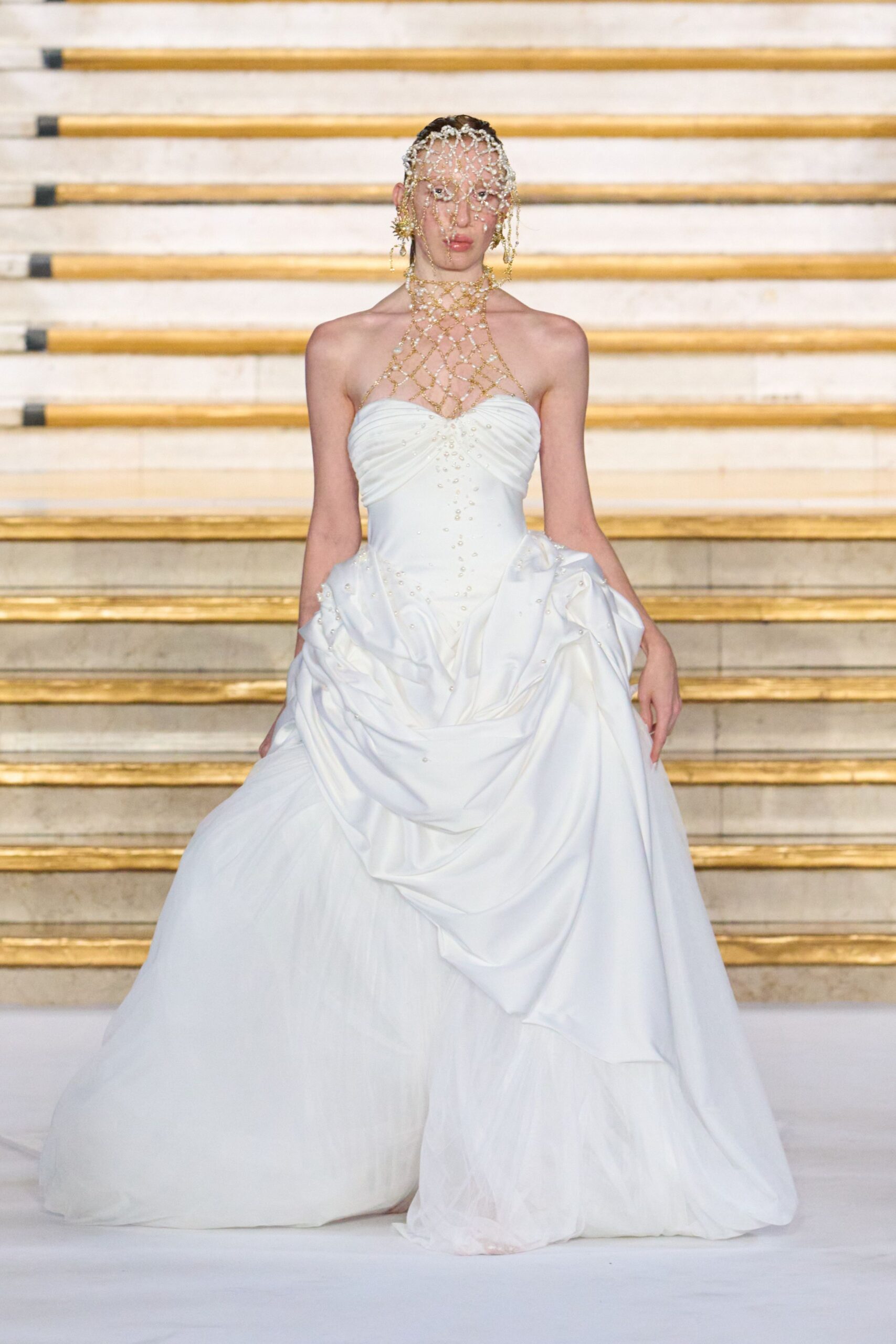 Pearl-strung net veil draped over the face from a close cap. Gold bead lattice climbing from throat to sternum. Spiky starburst drops at both ears. Ivory strapless satin gown, ruched sweetheart bodice and gathered overskirt over tulle. For the staircase descent after a wedding that happened in a museum.