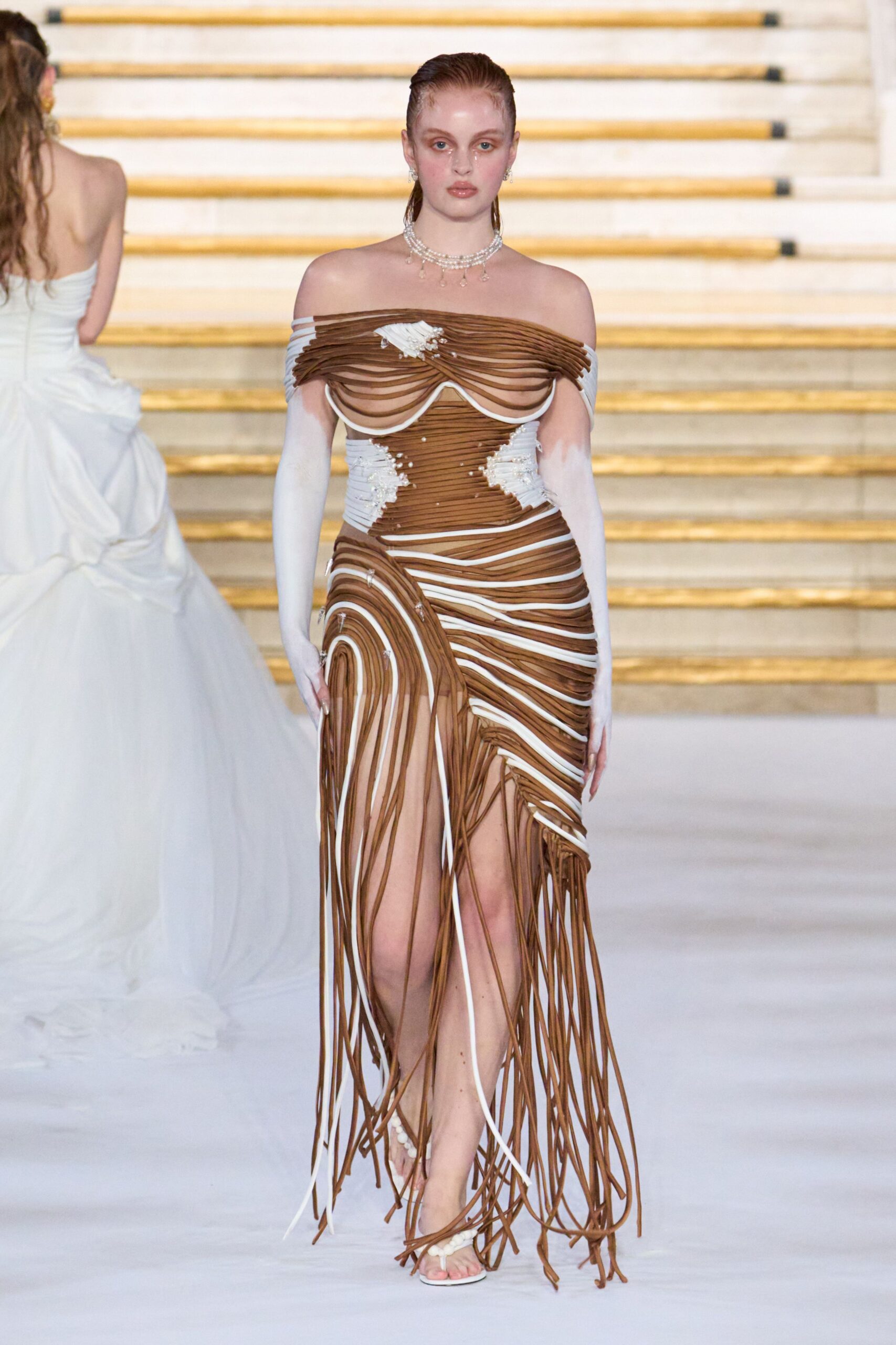 Tiered pearl strands with teardrop crystals at the throat. Off-shoulder gown built from cocoa and white cords, pearl clusters at the bust. Long white opera gloves fading sheer at the upper arm. White sandals with a beaded toe strap. Descending a marble staircase after the wedding cake has been cut.