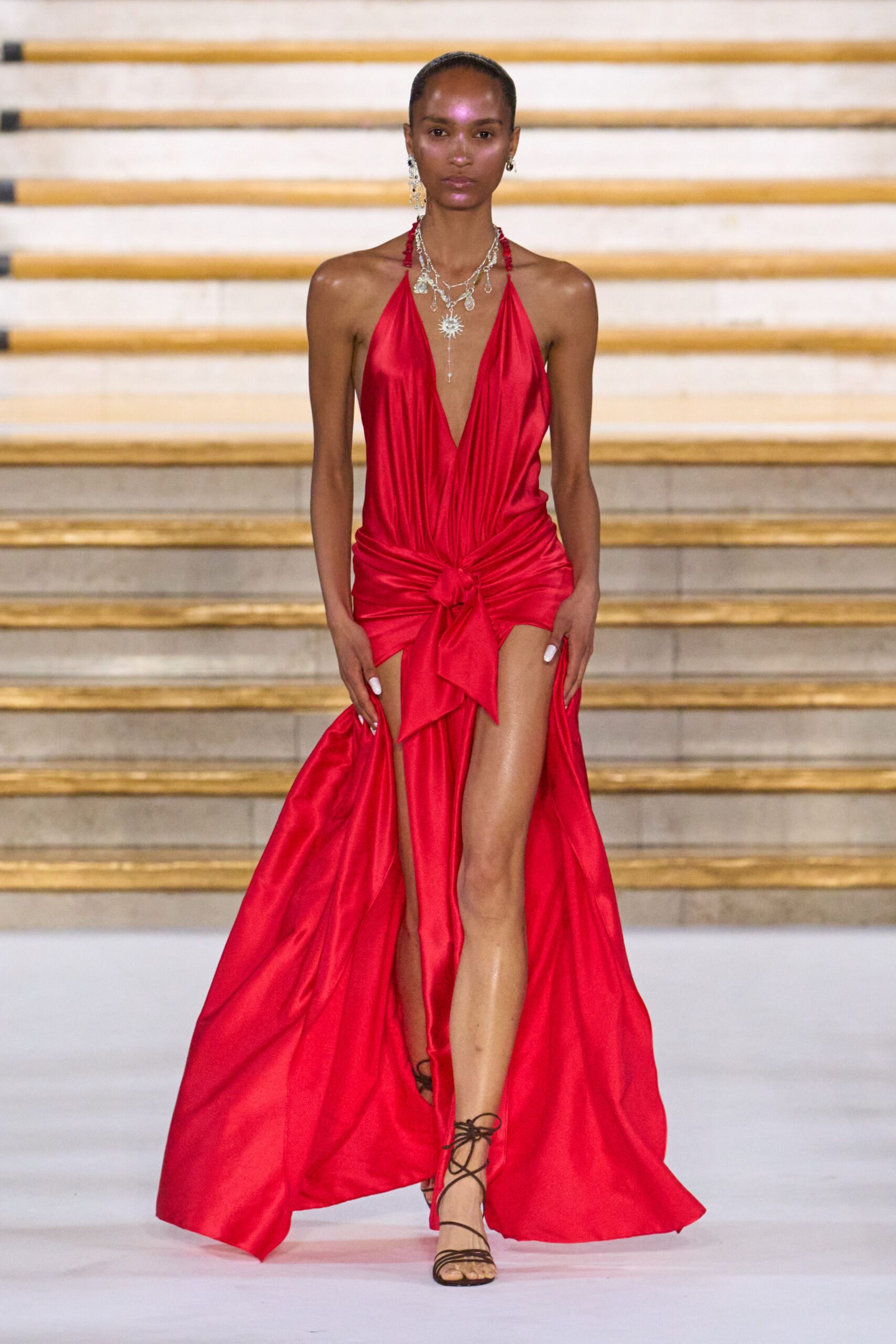 Layered silver charms and crystals, one sunburst dropping to the sternum. Scarlet satin halter gown, plunging to the waist with a knotted hip drape. Dark lace-up sandals, cords winding fine around the ankles. Crossing a hotel ballroom after midnight, hem sweeping the waxed floor.