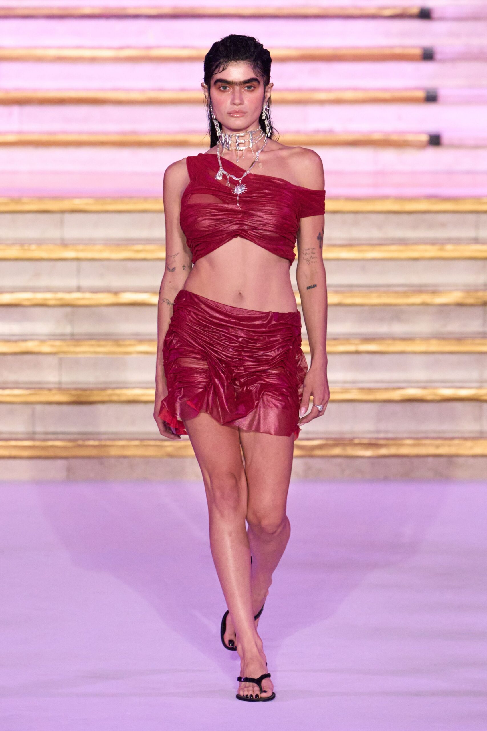 Wine-red mesh crop top, ruched tight with one shoulder fallen low. Matching micro mini, crinkled and gathered with a shredded side panel. Layered silver chains with a square-link choker and tiny sun charm. Black thong sandals with fuzzy straps. After midnight on hotel steps still warm from the day.