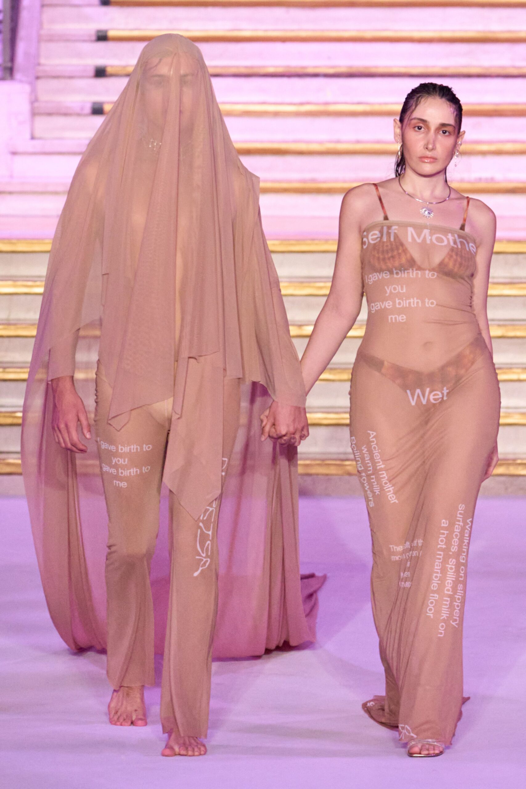 Floor-length nude mesh veil, draped over the head and face. Second-skin nude jersey printed with scattered white text, feet left bare. Holding hands on a marble staircase after everyone else has gone home.