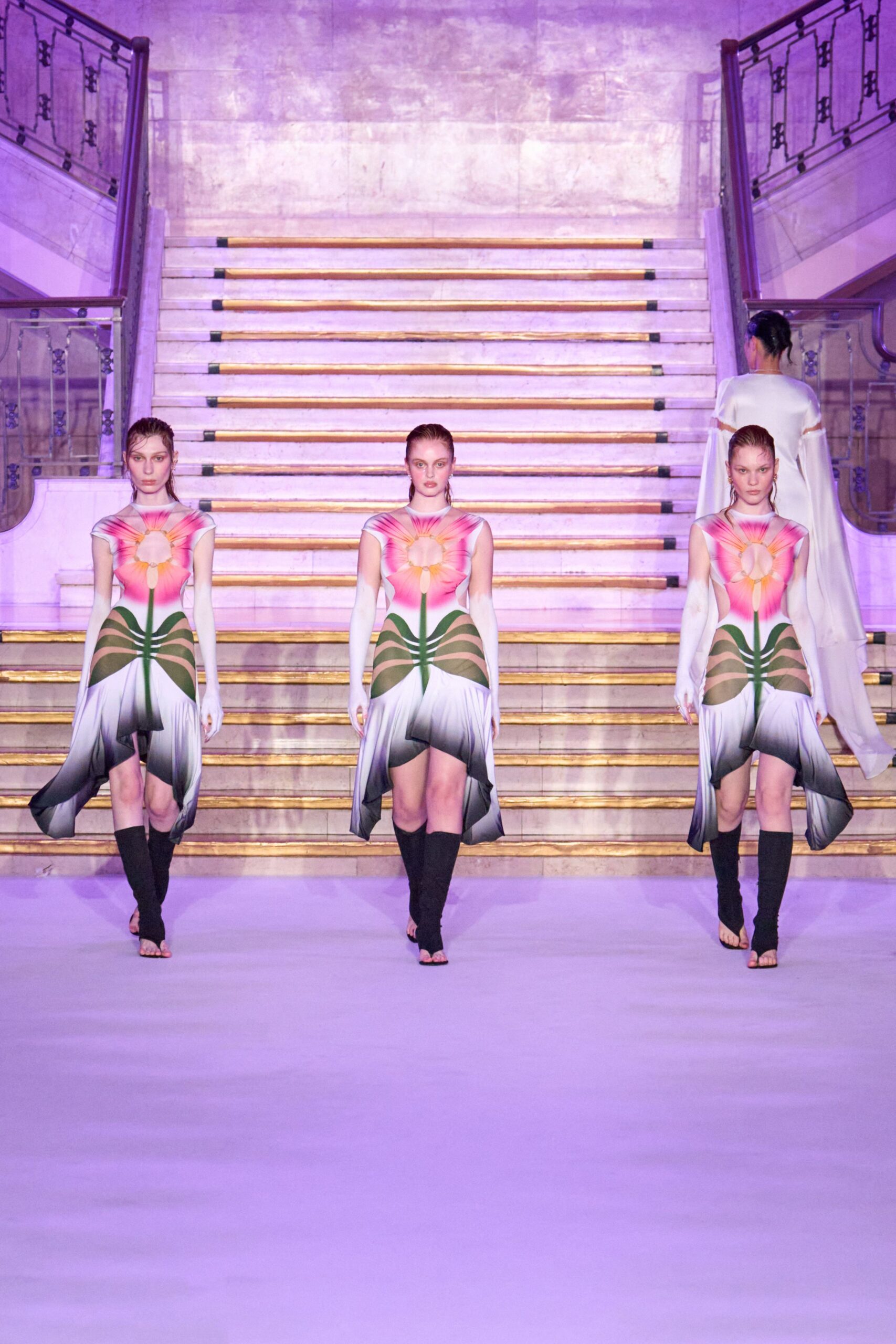 Body-skimming dress printed as a pink flower, with oval cutouts through the torso. Long white opera gloves disappearing into the cap sleeves. Black knit stirrup leg warmers, toes left bare. Descending a grand stair under lilac light, dressed for a masquerade in a conservatory.