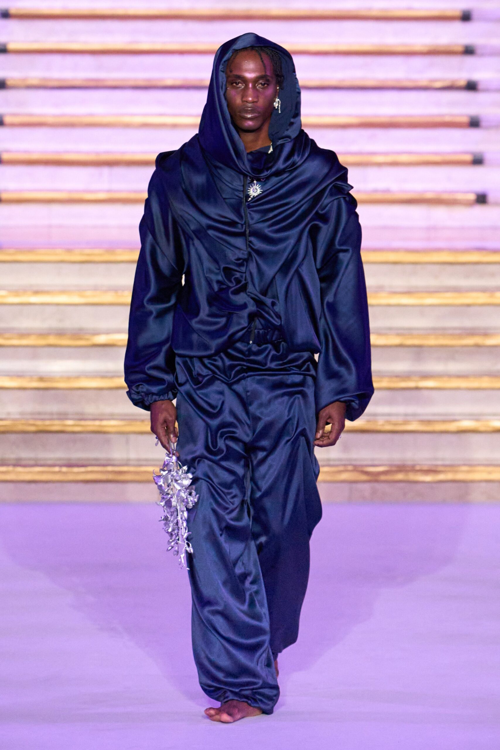 Midnight satin hooded blouse, cowl neck collapsing into gathered seams. Matching satin trousers, elastic waist and ankles puckered into shine. Sunburst pendant caught at the throat. Single silver dangling earring. Metallic lilac flower spray gripped by the stems. Bare feet on cold tile after carrying wedding flowers upstairs.