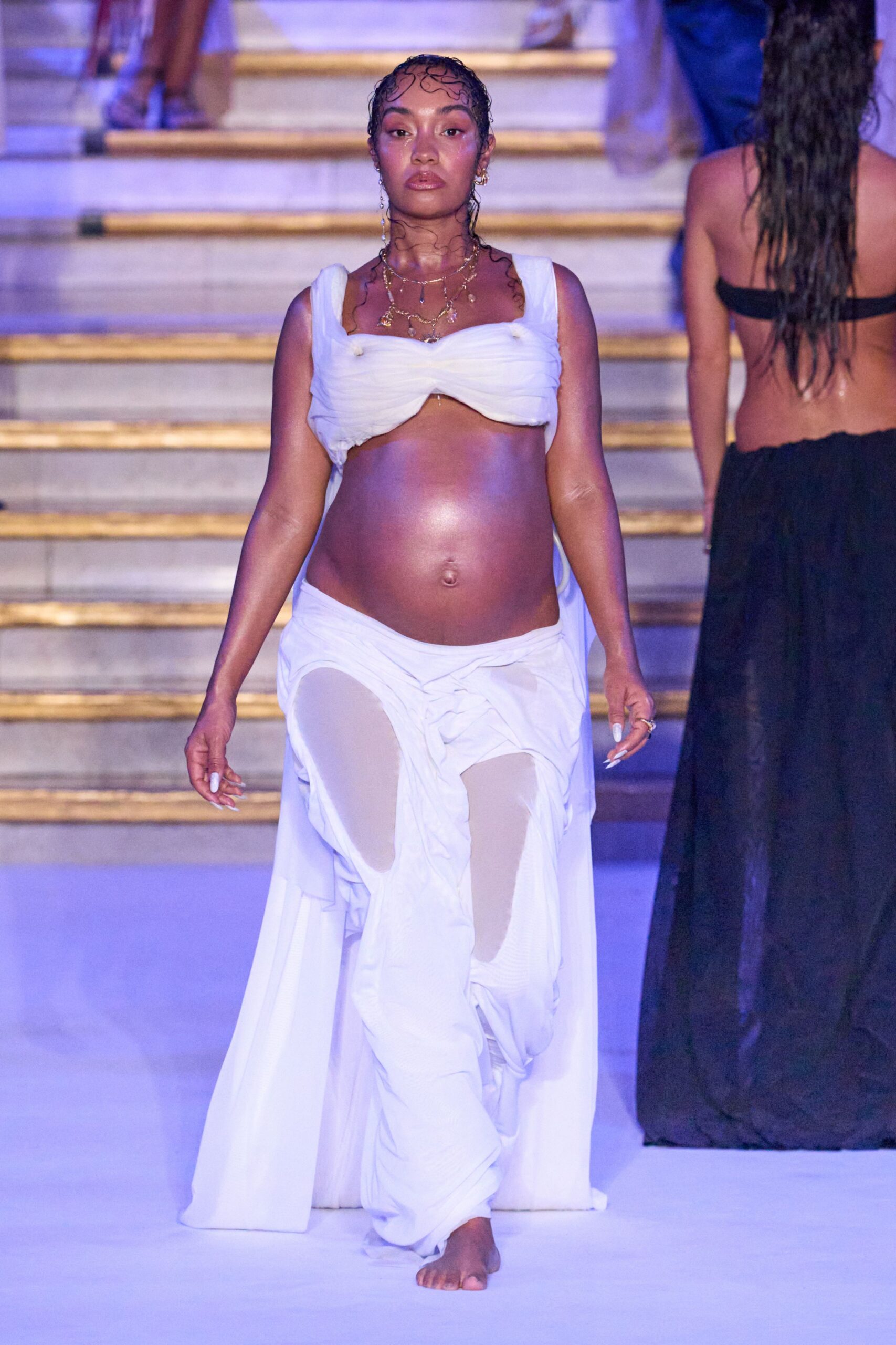 White ruched bandeau top with thick straps and a pinched center seam. Low-slung white draped skirt, split open over sheer stockinged legs. Layered gold chains with tiny shell-like drops at the collarbone. Walking barefoot down seaside steps after a swim that ran past sunset.