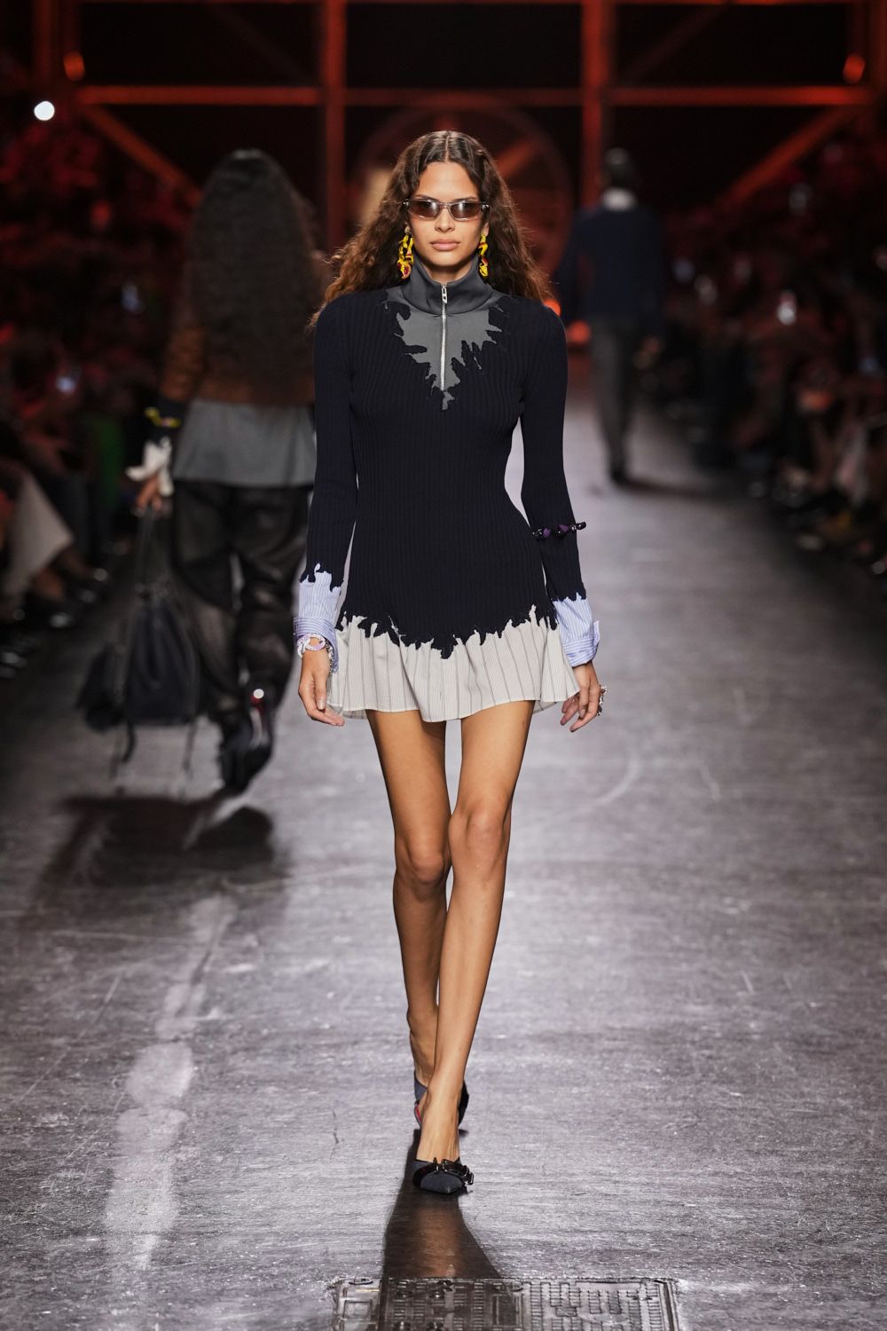 Black ribbed knit mini dress with trompe-l'oeil white shirt collar and pleated hem. Yellow and red dangling figures. Narrow smoked lenses. Black pointed flats with a small bow across the vamp. Walking to a lunch meeting with rain still drying on the pavement.