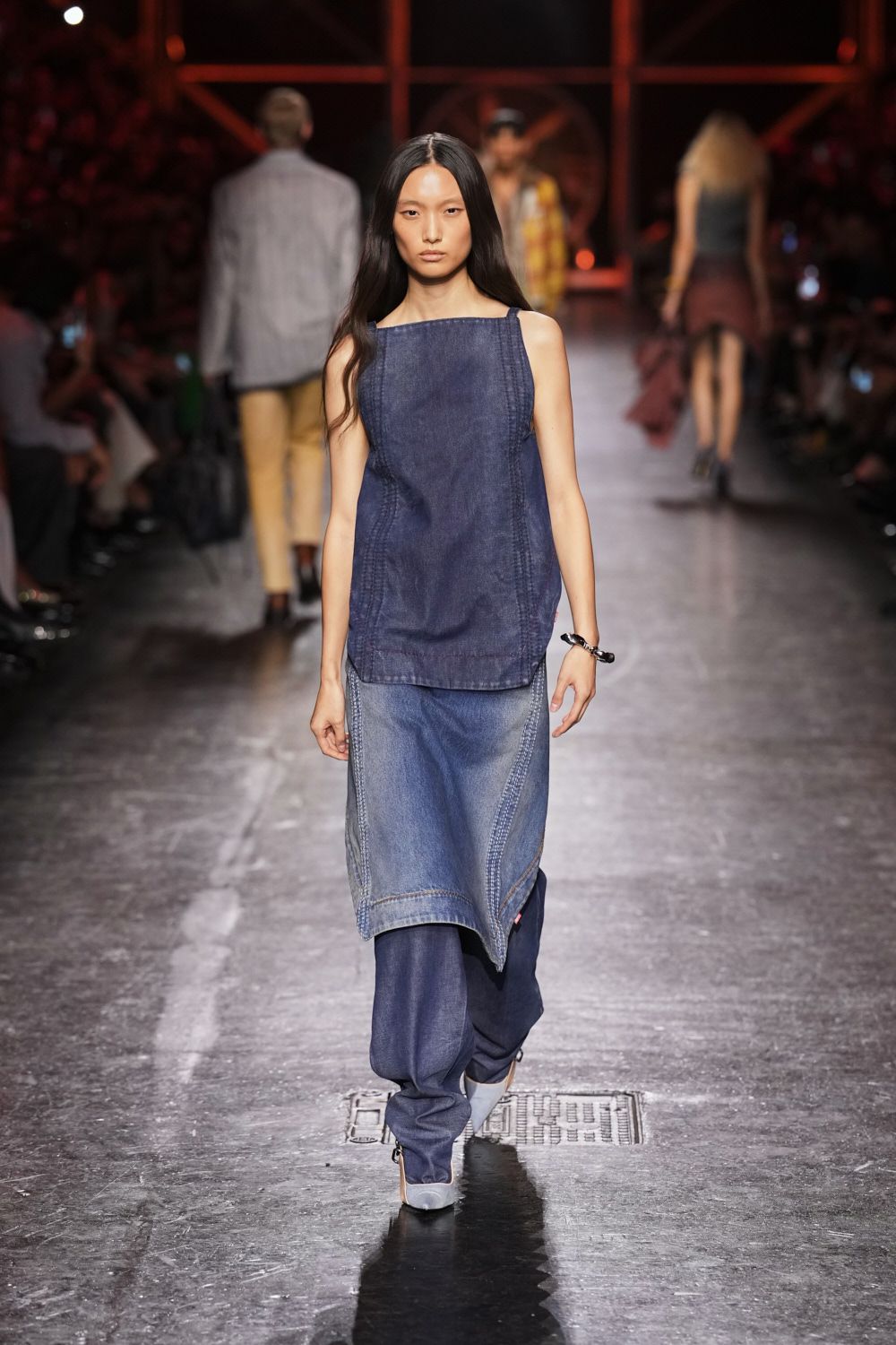 Dark indigo denim tank with a square neckline and doubled side seams. Faded blue denim apron skirt, curved panels ending in a pointed hem. Slouchy dark-wash jeans pooling over the shoes. Twisted black bangle at one wrist. White square-toe heels. Crossing the fairground after midnight with dust still stuck to your cuffs.